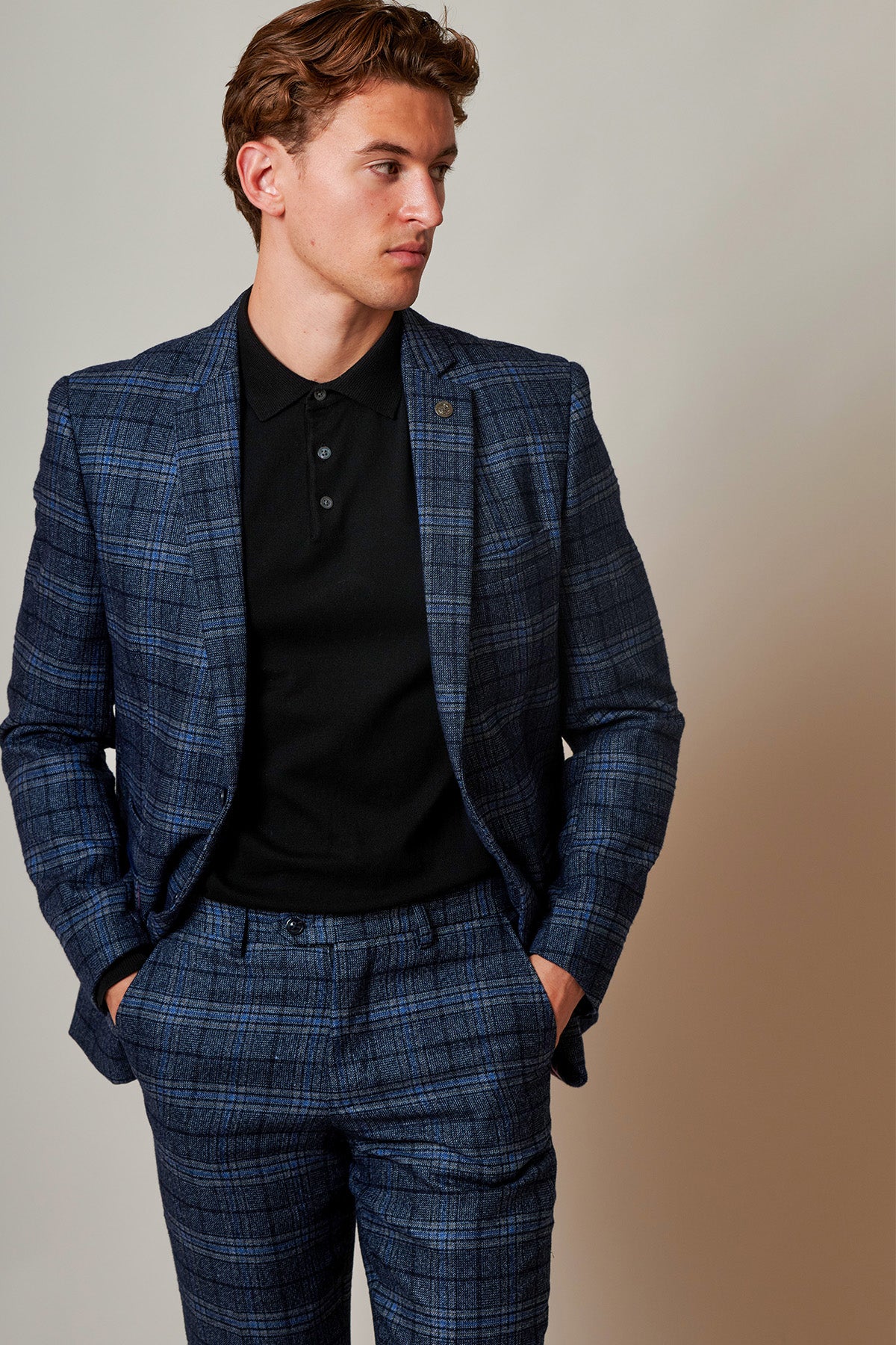 A man in the ANDY - Blue Check Two Piece Suit by Marc Darcy, paired with a black polo shirt, stands with hands in pockets and looks left against a plain, light-colored background.
