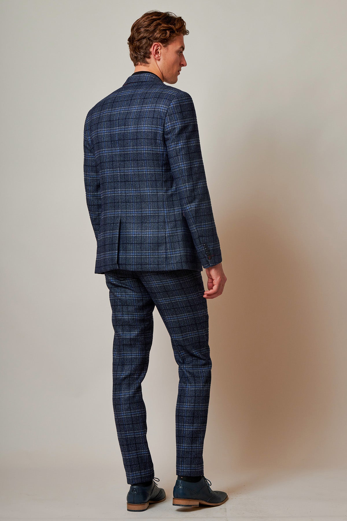 A man stands with his back to the camera, wearing the Marc Darcy ANDY Blue Check Two Piece Suit and dark blue shoes against a plain, light background.