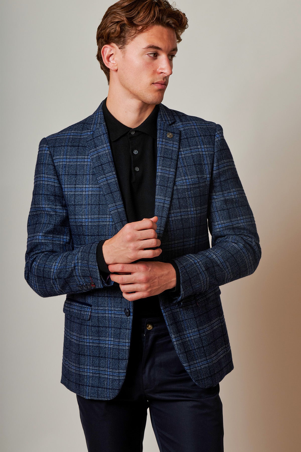 A man wearing the Marc Darcy ANDY - Blue Check Blazer in a slim fit over a black shirt and dark pants stands against a plain light background, looking to his left and adjusting his blazer.