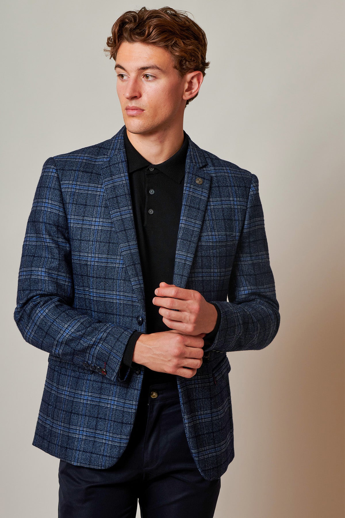 A young man in the Marc Darcy ANDY Blue Check Blazer stands against a plain background, wearing it over a black polo shirt, adjusting his jacket with one hand and looking to the side.