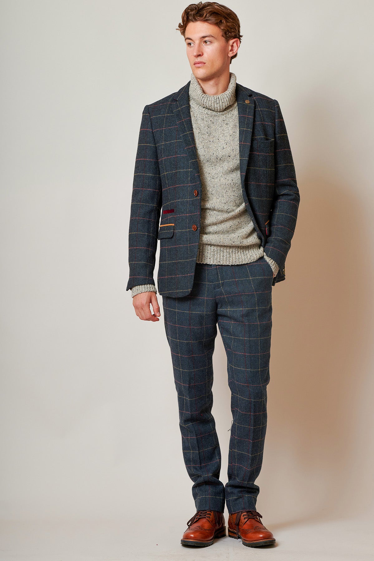 A man with short brown hair stands confidently against a plain background, wearing the marcdarcy ETON Navy Blue Tweed Check Blazer, a light grey knit sweater, and brown leather shoes, with one hand in his pocket.