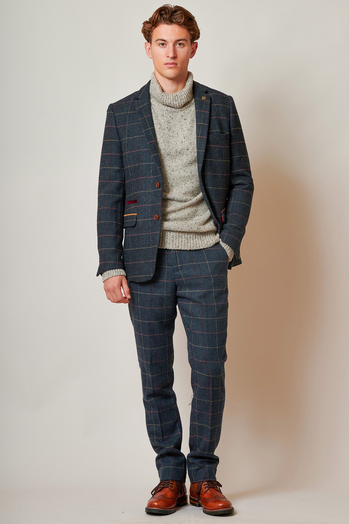 A young man with short brown hair and a neutral expression stands against a plain background, wearing the marcdarcy ETON - Navy Blue Tweed Check Blazer over a grey turtleneck sweater and paired with brown leather shoes.