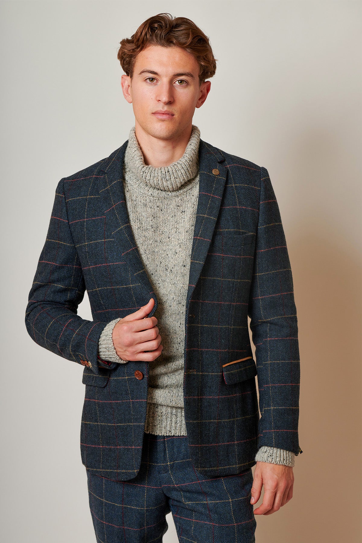 A young man with wavy brown hair wears a gray turtleneck and the marcdarcy ETON Navy Blue Tweed Check Blazer with matching trousers, standing confidently against a light background in smart casual style.