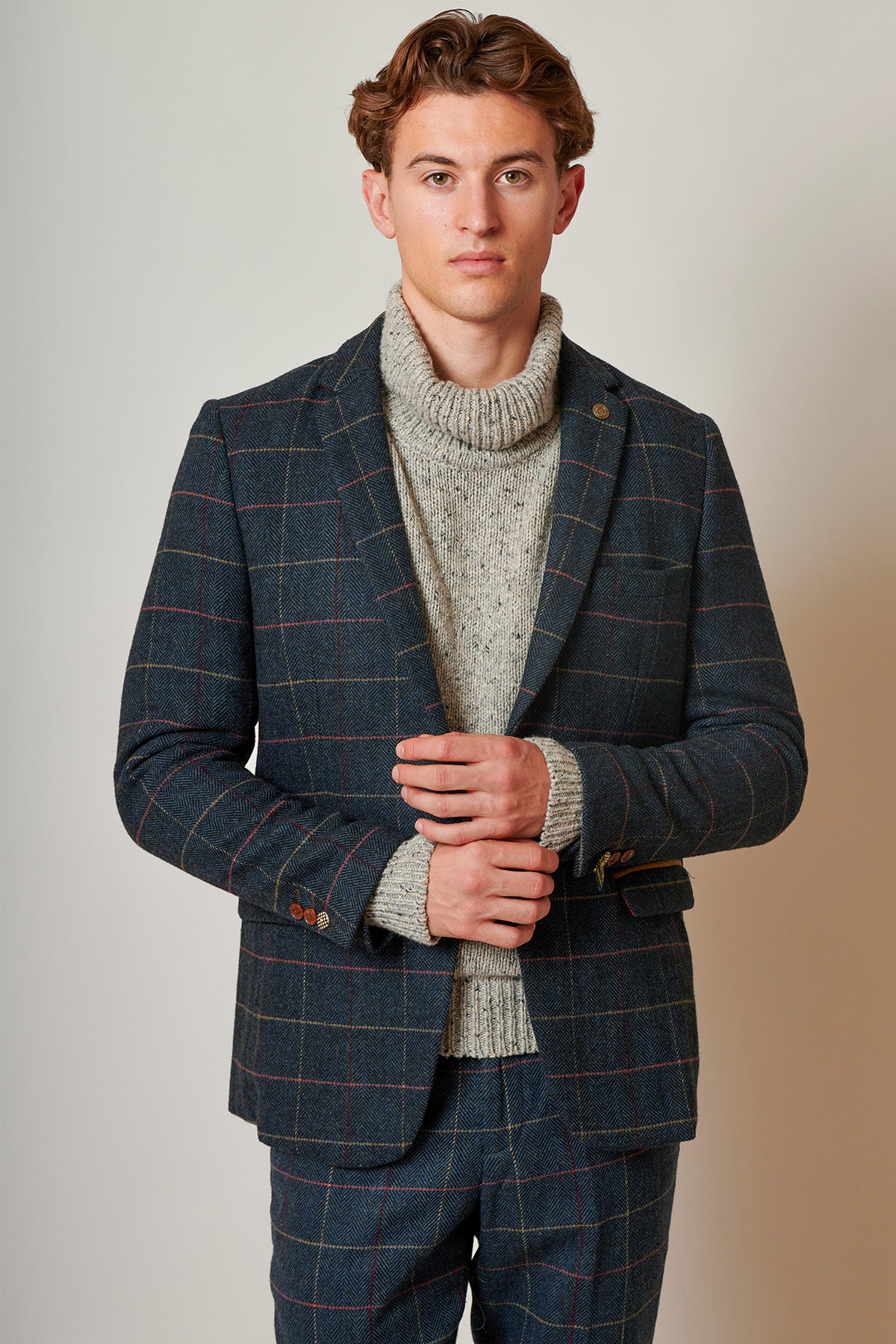 A young man stands against a neutral background, wearing the marcdarcy ETON Navy Blue Tweed Check Blazer over a grey chunky knit turtleneck, embodying smart casual style with a neutral expression and hands clasped.