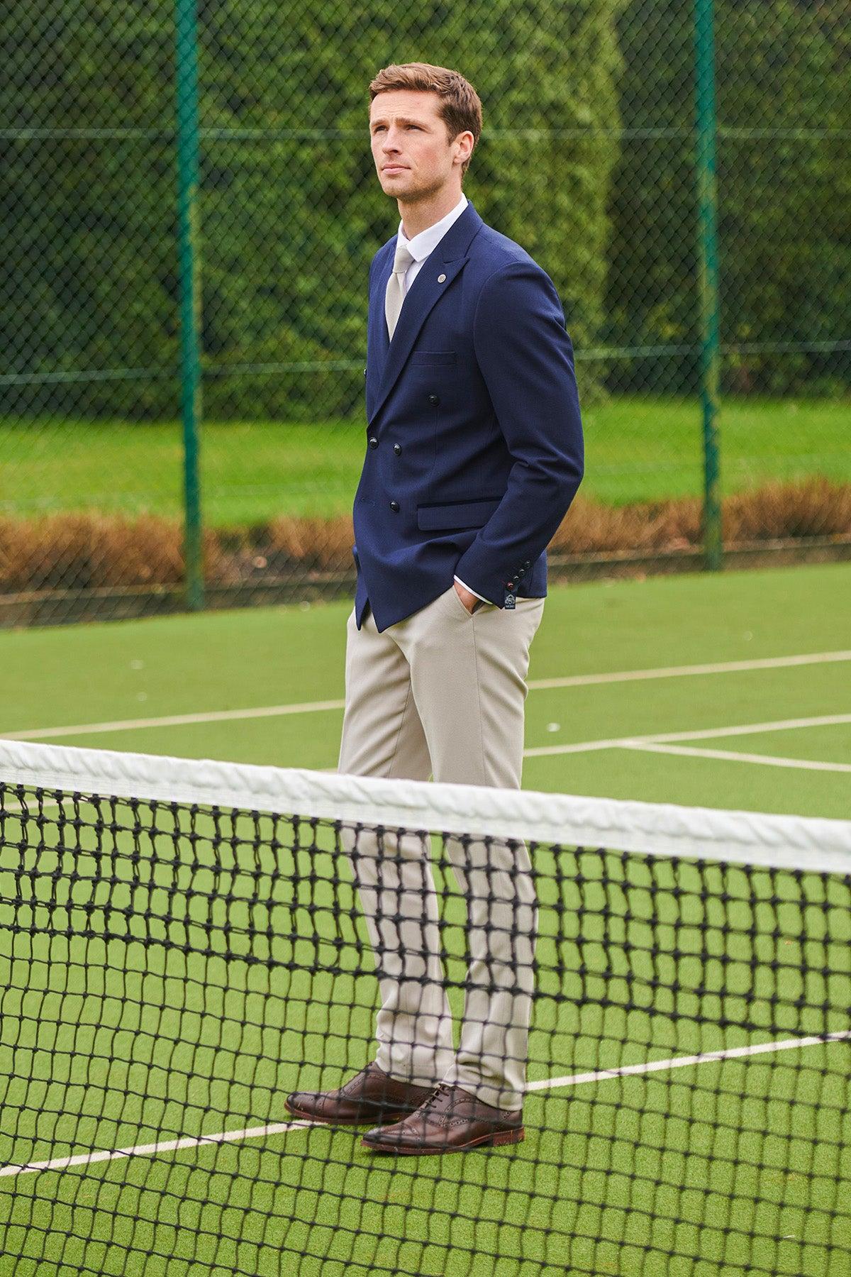 A man in the Marc Darcy BROMLEY navy double-breasted blazer and HM5 Stone trousers stands with hands in pockets on a tennis court, gazing into the distance. Trees and fencing frame the modern tailoring-inspired scene.