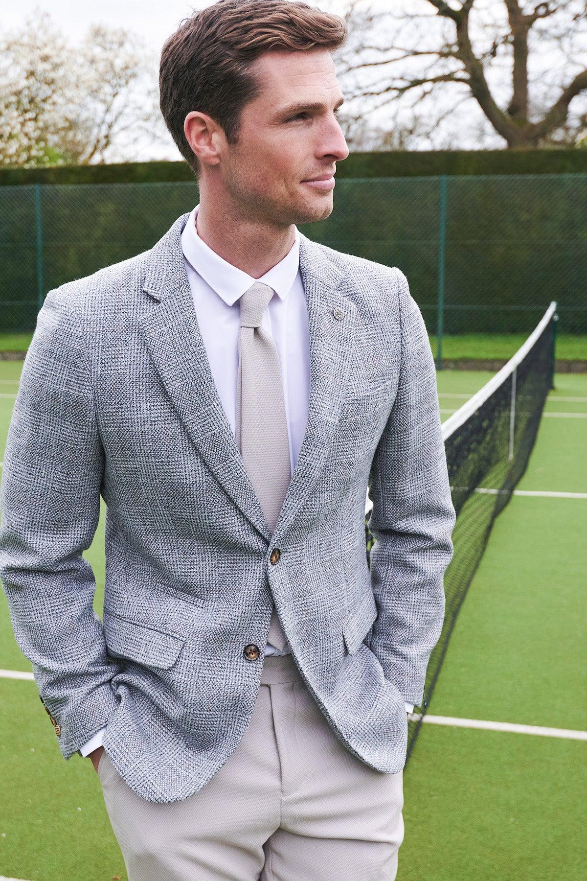 A man wearing the marcdarcy KRIS Silver Grey Tweed Blazer with HM5 Stone Trousers stands on a tennis court, hands in pockets and looking aside, with the net and green court visible behind him.