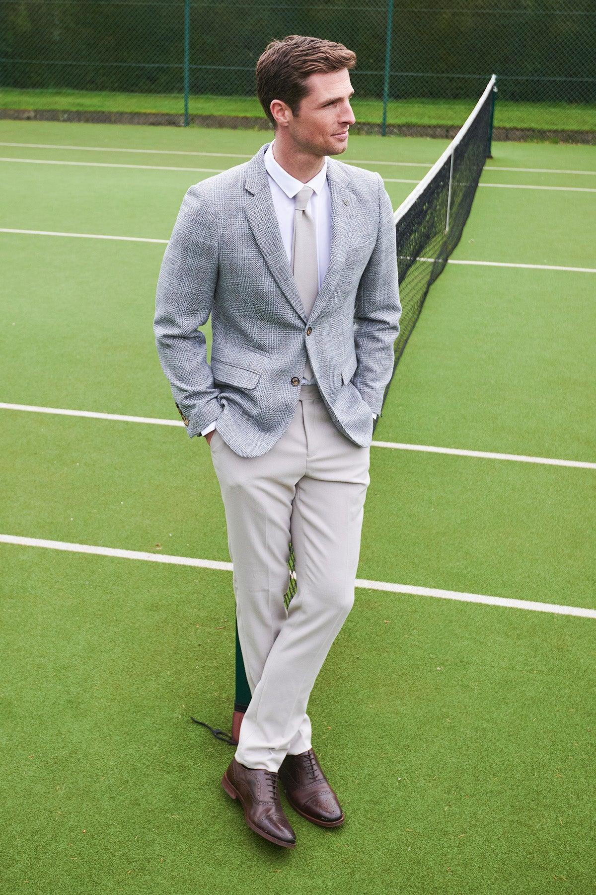 A man wears the marcdarcy KRIS Silver Grey Tweed Blazer with HM5 Stone Trousers while standing on a tennis court.