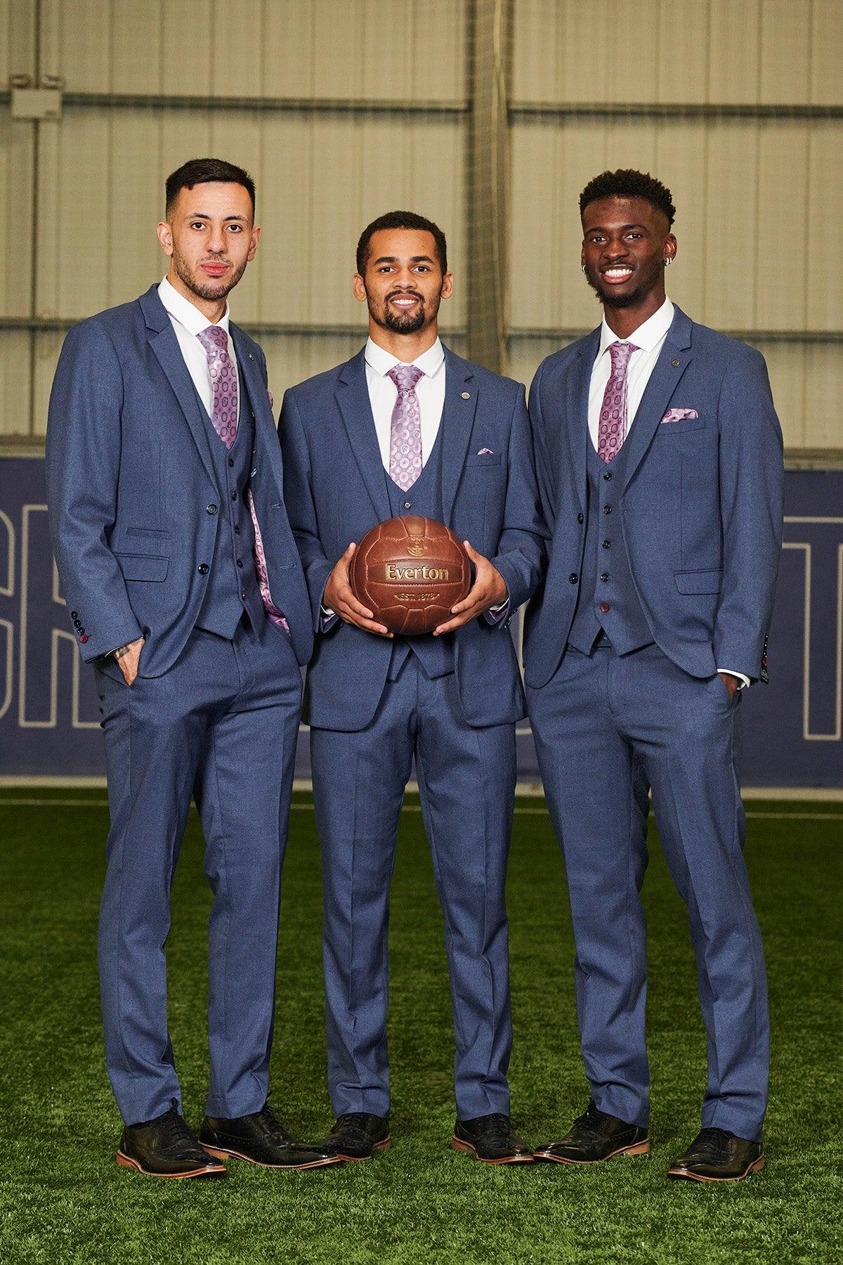 Three men in matching The Everton Collection | ASHLEY Navy Blue Three Piece Suits by Marc Darcy stand on a green field. The man in the middle holds a vintage brown football.