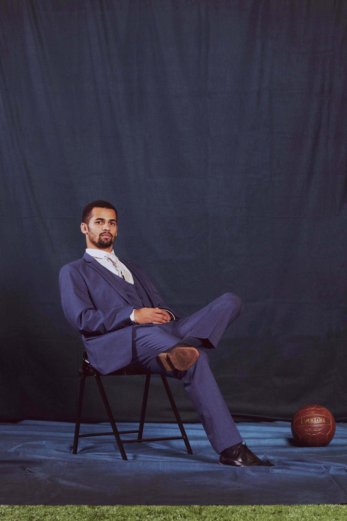 Iliman Ndiaye wears the Marc Darcy ASHLEY Navy Blue Three Piece Suit from The Everton Collection, sitting on a black chair with crossed legs as he looks at the camera. A vintage soccer ball sits nearby.