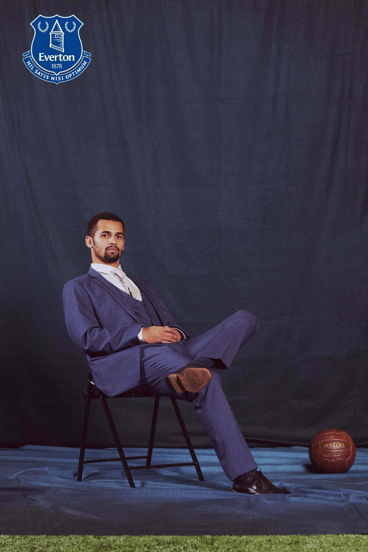 Iliman Ndiaye, in a Marc Darcy ASHLEY Navy Blue Three Piece Suit from The Everton Collection, sits with legs crossed before a navy backdrop featuring the Everton FC crest and collection items in the top left.