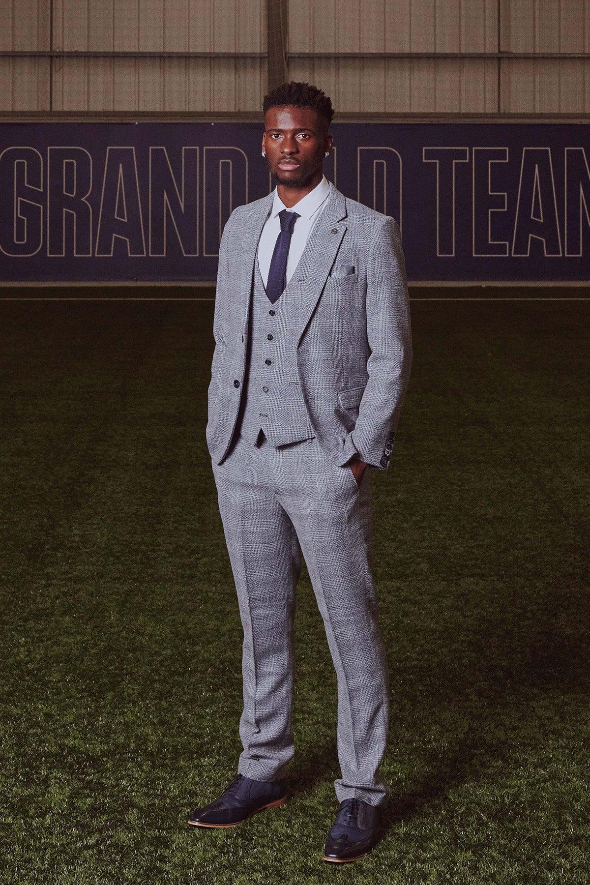 Tim Iroegbunam stands on a green turf field wearing marcdarcy’s The Everton Collection KRIS Sky Blue Tweed Suit. A navy banner reading GRAND TEAM is seen in the background.