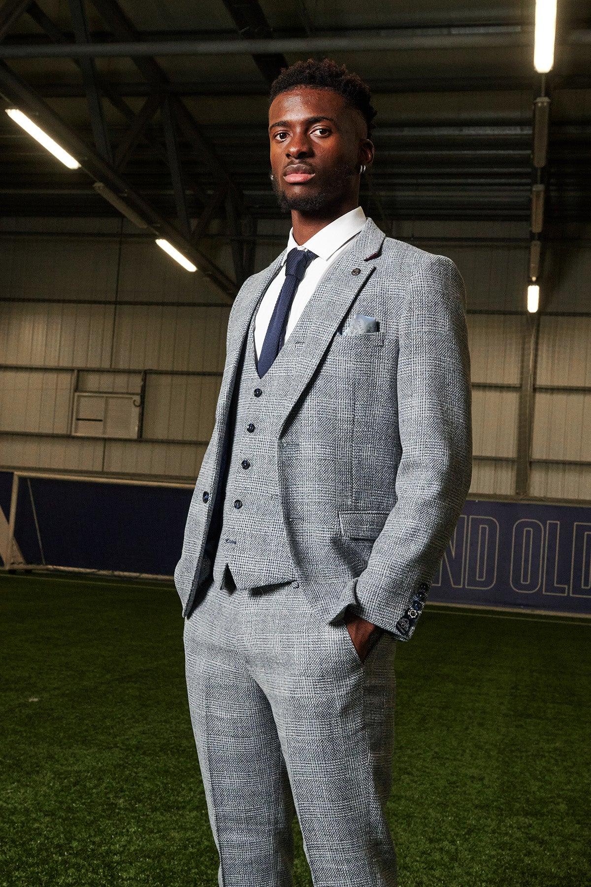 A Black man stands in a green field wearing the marcdarcy Everton Collection, featuring Tim Iroegbunam in the KRIS Sky Blue Tweed Suit, his hands in his pockets and exuding classic elegance.