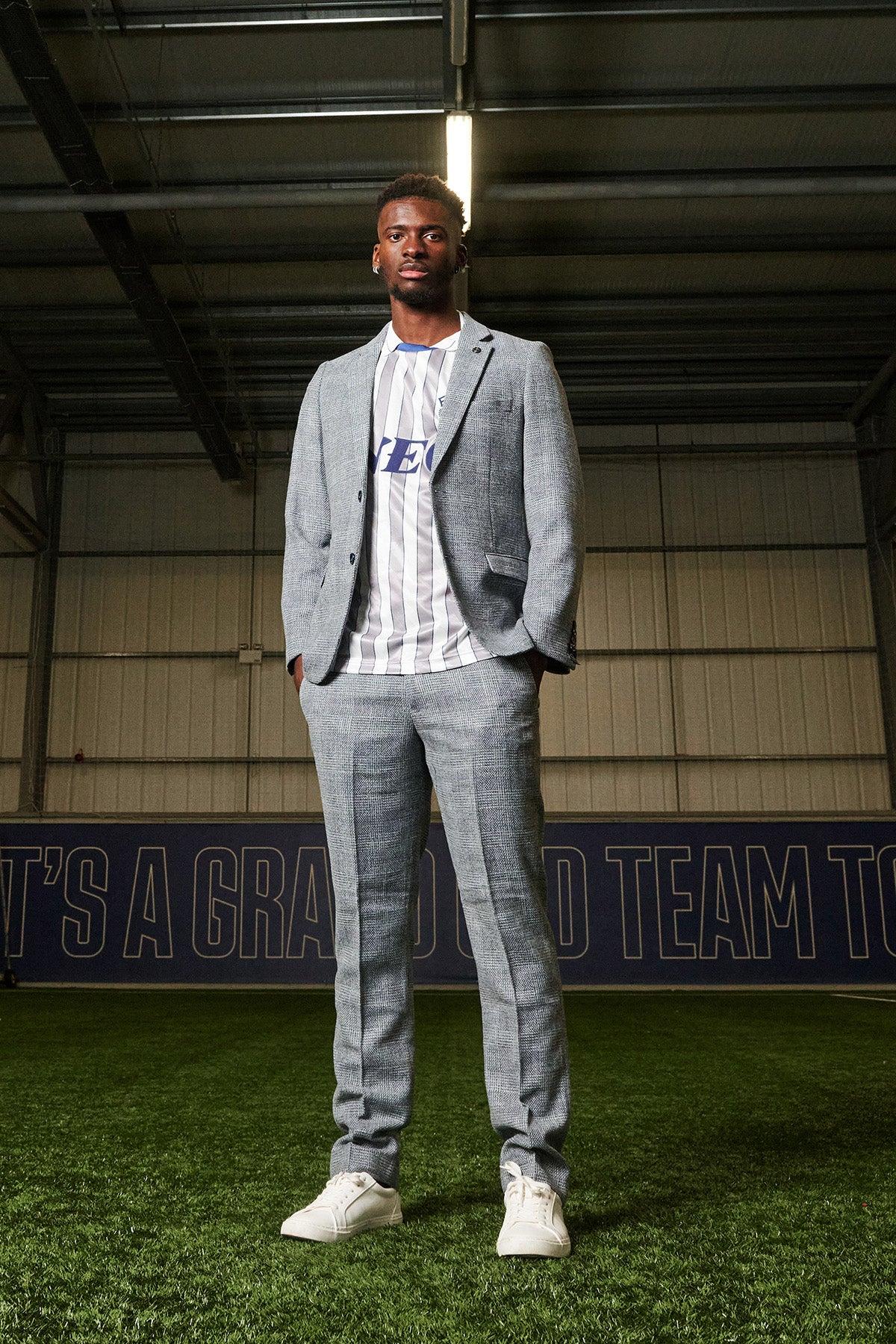 Tim Iroegbunam stands on indoor turf in the marcdarcy KRIS Sky Blue Tweed Suit from The Everton Collection, over a striped sports jersey and white sneakers, confidently posing with a metal roof and blue banner in the background.