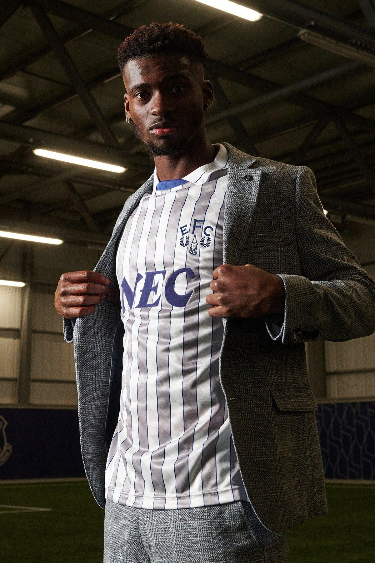 Tim Iroegbunam stands indoors on artificial turf, revealing an Everton football jersey beneath his marcdarcy KRIS Sky Blue Tweed Suit from The Everton Collection.