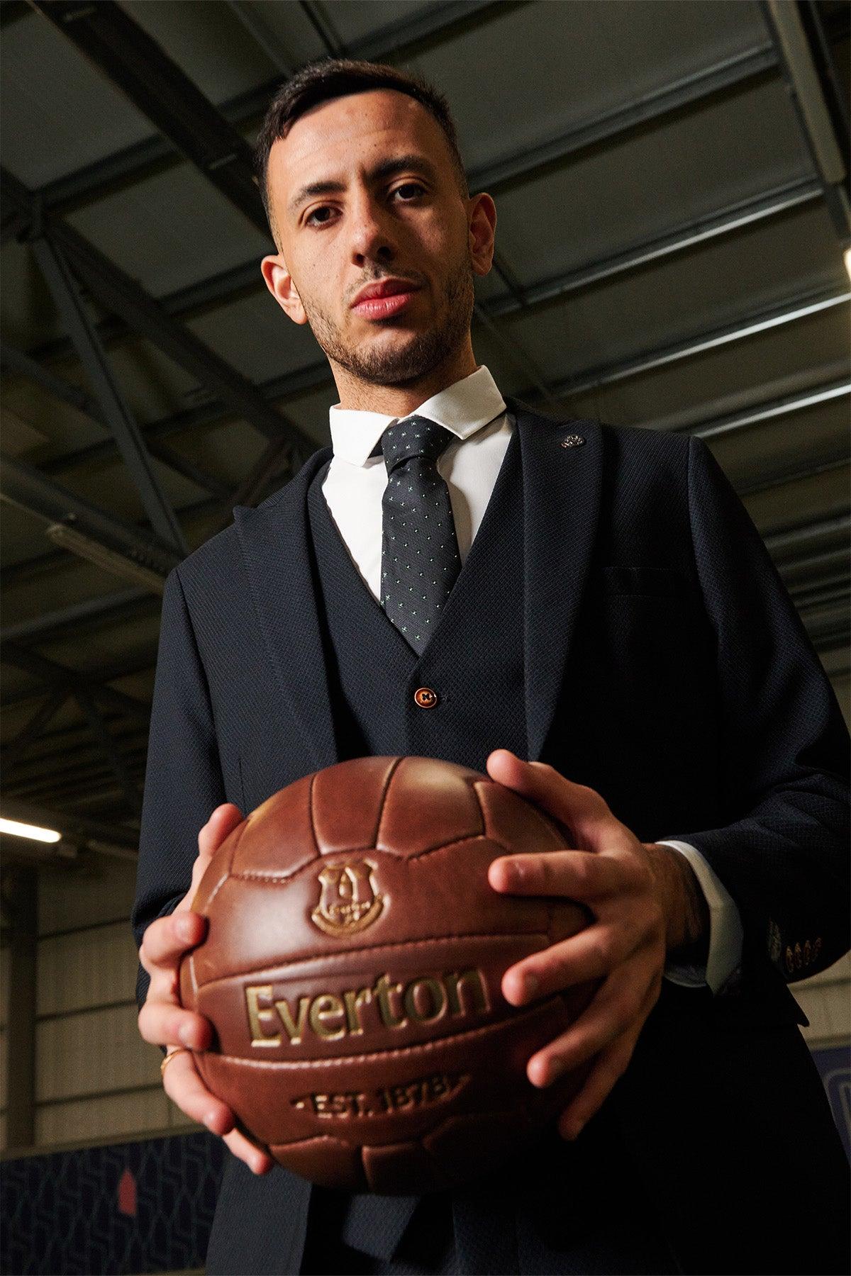 Dwight McNeil stands indoors under industrial lights, wearing a marcdarcy Max Navy Suit from The Everton Collection and holds a vintage brown leather football embossed with Everton and EST. 1878.