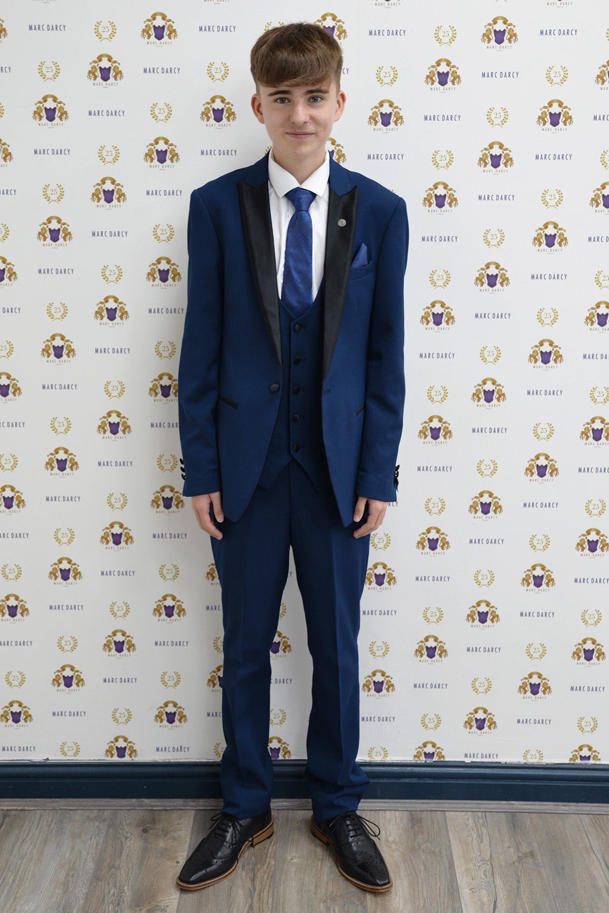 Charlie Wrenshall as Liam Connor wears the marcdarcy DALTON Royal Blue Tux Suit—a slim fit tux with peak lapels—paired with a white shirt and blue tie, smiling before a white backdrop with gold and blue logos on a wooden floor.