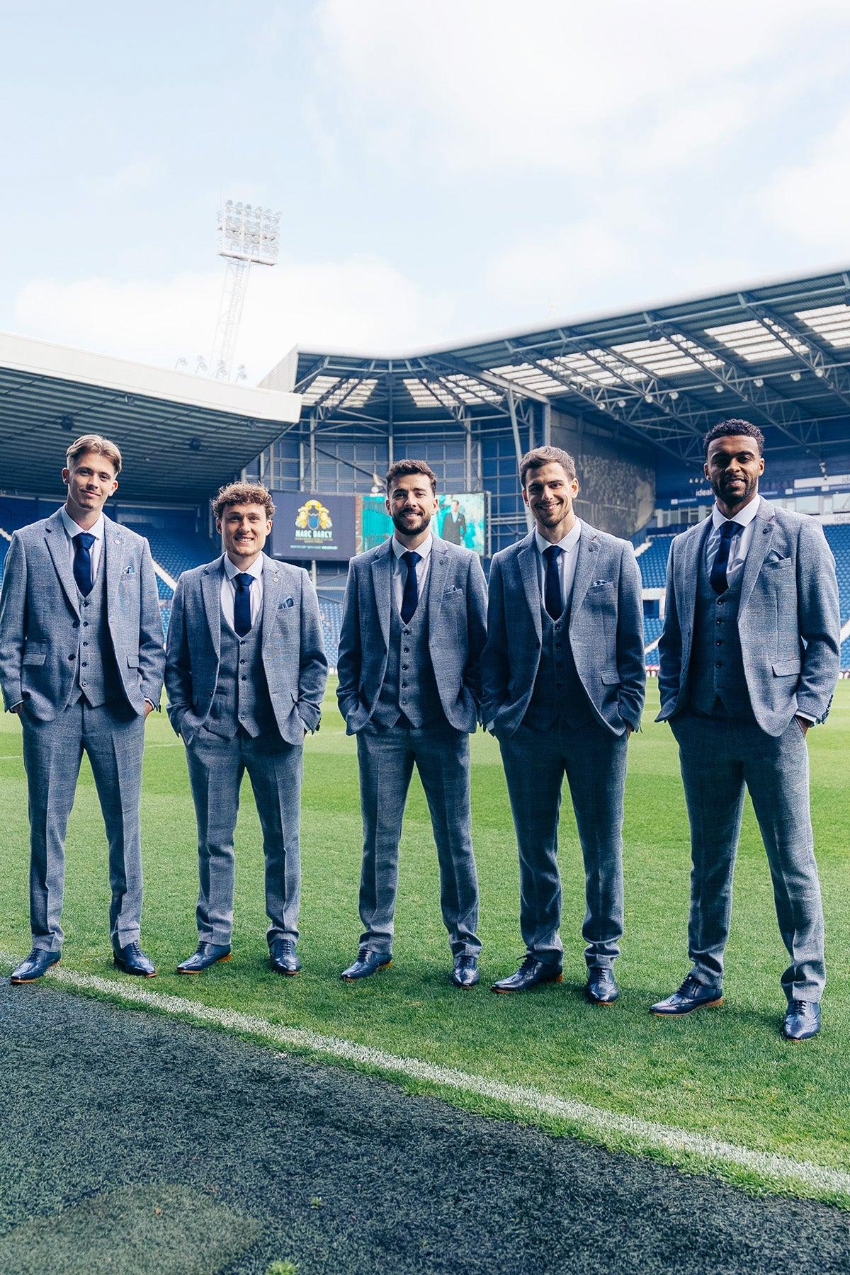 Five men in matching marcdarcy Official West Bromwich Albion F.C. Club KRIS Sky Blue Tweed Suits and navy ties stand on a soccer field in front of stadium seating.