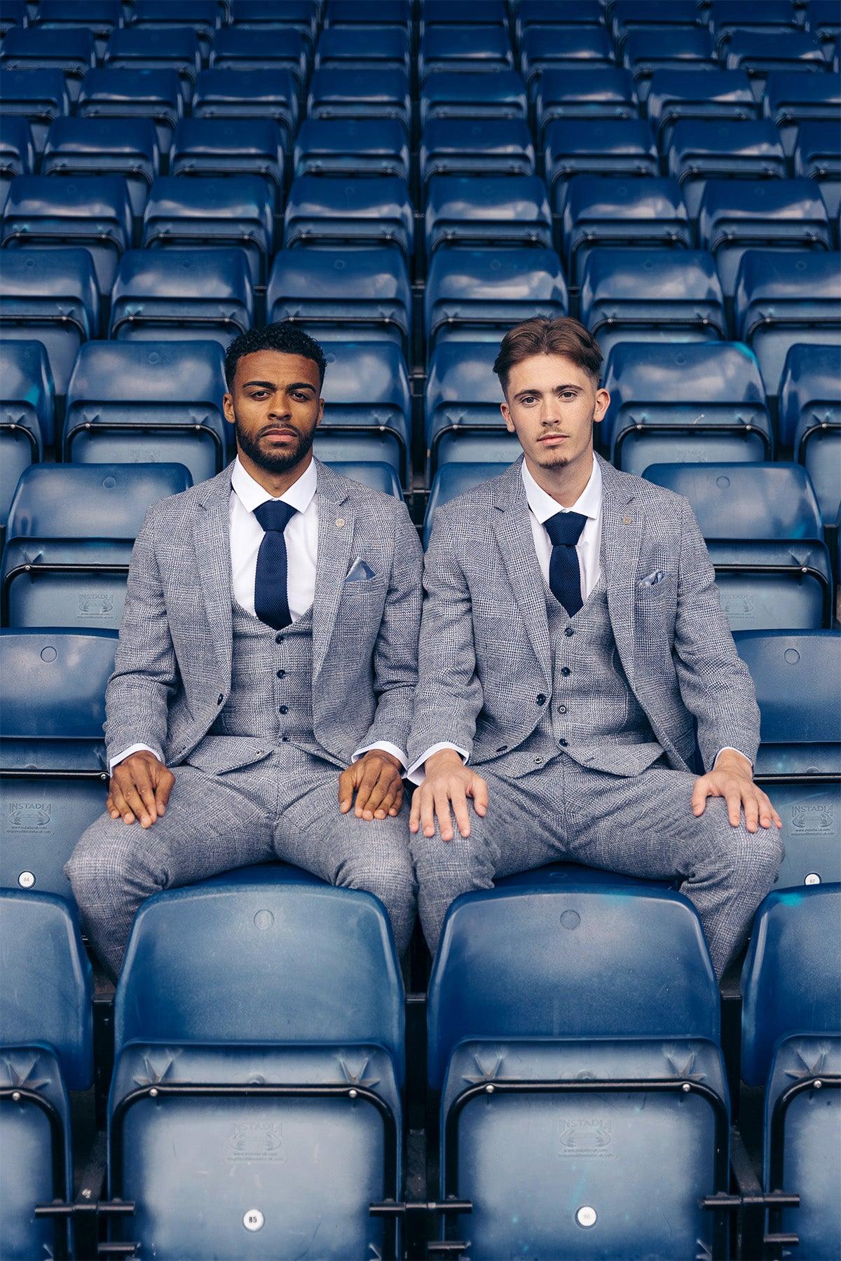 Two men in matching marcdarcy Official West Bromwich Albion F.C. Club Suits (KRIS Sky Blue Tweed) sit side by side in empty blue stadium seats, wearing dark ties and looking seriously at the camera.