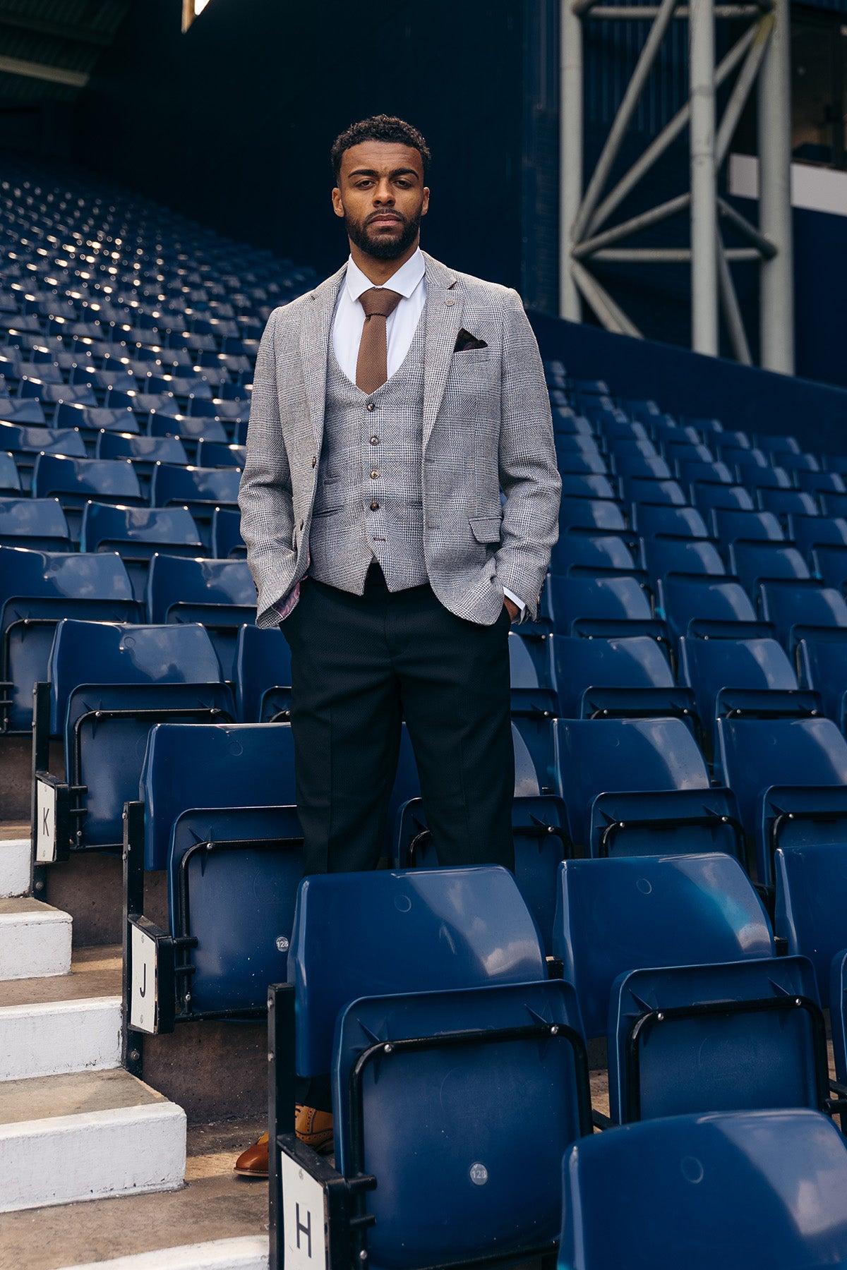A man wears the marcdarcy West Bromwich Albion F.C. Collection: KRIS Silver Grey Blazer and Max Black Trousers, as seen on Darnell Furlong, standing in empty stadium seating with a white shirt and brown tie.