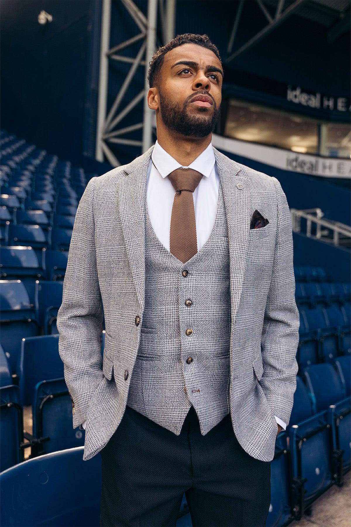 A man in the marcdarcy West Bromwich Albion F.C. Collection wears the KRIS Silver Grey Blazer and Max Black Trousers, as seen on Darnell Furlong, standing confidently in an empty stadium with blue seats, beard neatly groomed.
