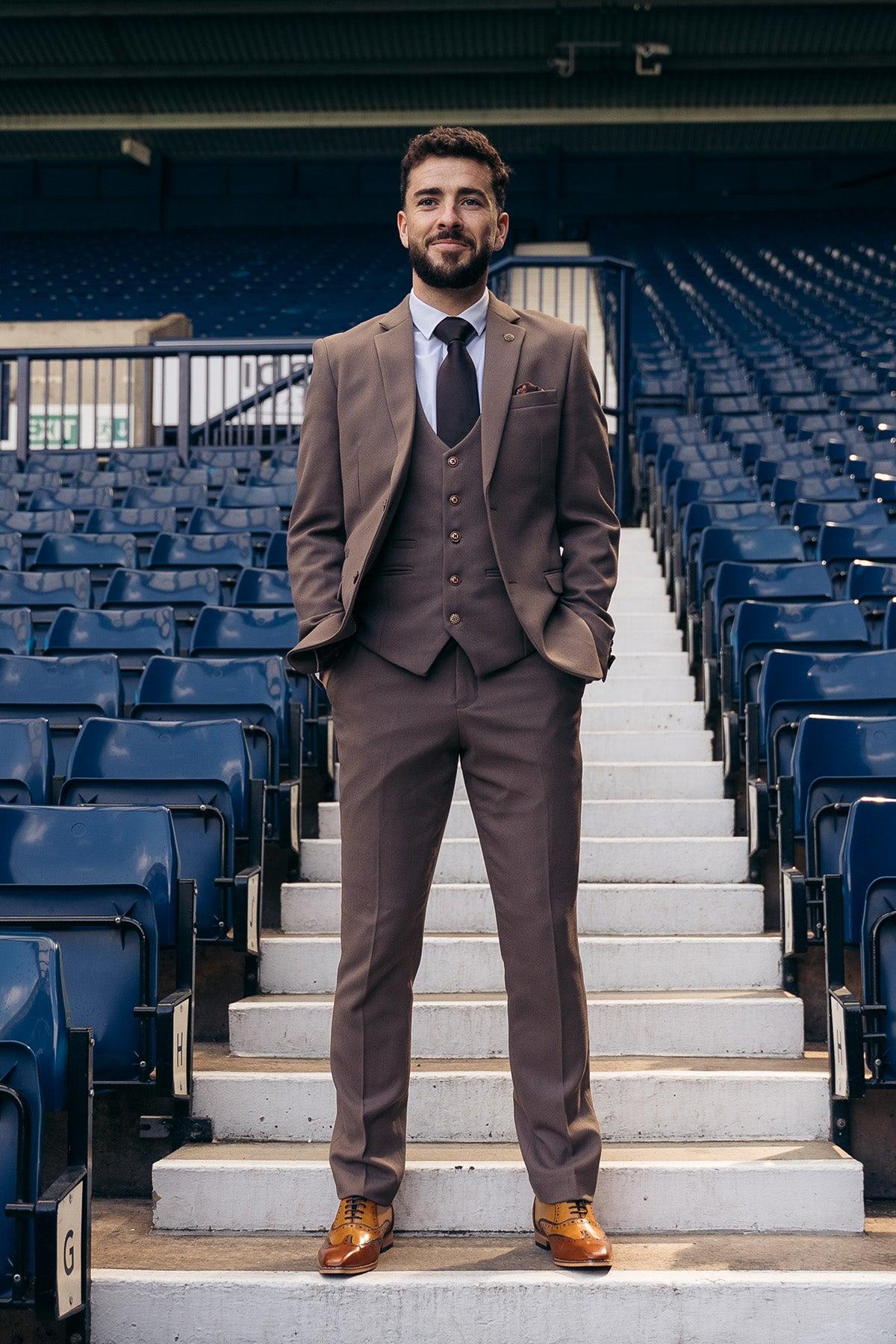 A man wears the Marc Darcy West Bromwich Albion F.C. Collection HM5 Tan Tailored Suit, as seen on Michael Johnston, standing confidently on stadium steps before rows of empty blue seats.