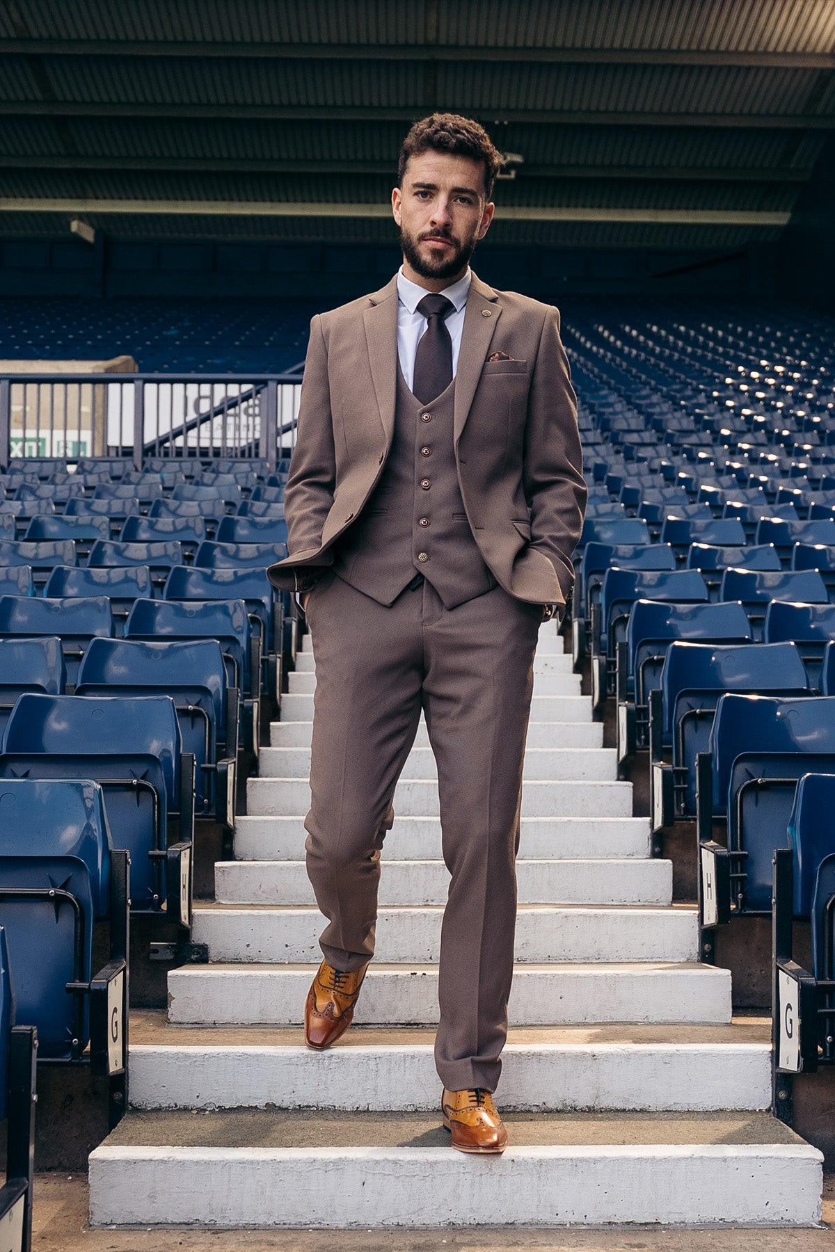 A man wearing the Marc Darcy HM5 Tan Tailored Suit from The West Bromwich Albion F.C. Collection stands confidently on a stadium staircase among empty blue seats, hands in pockets, looking directly at the camera.