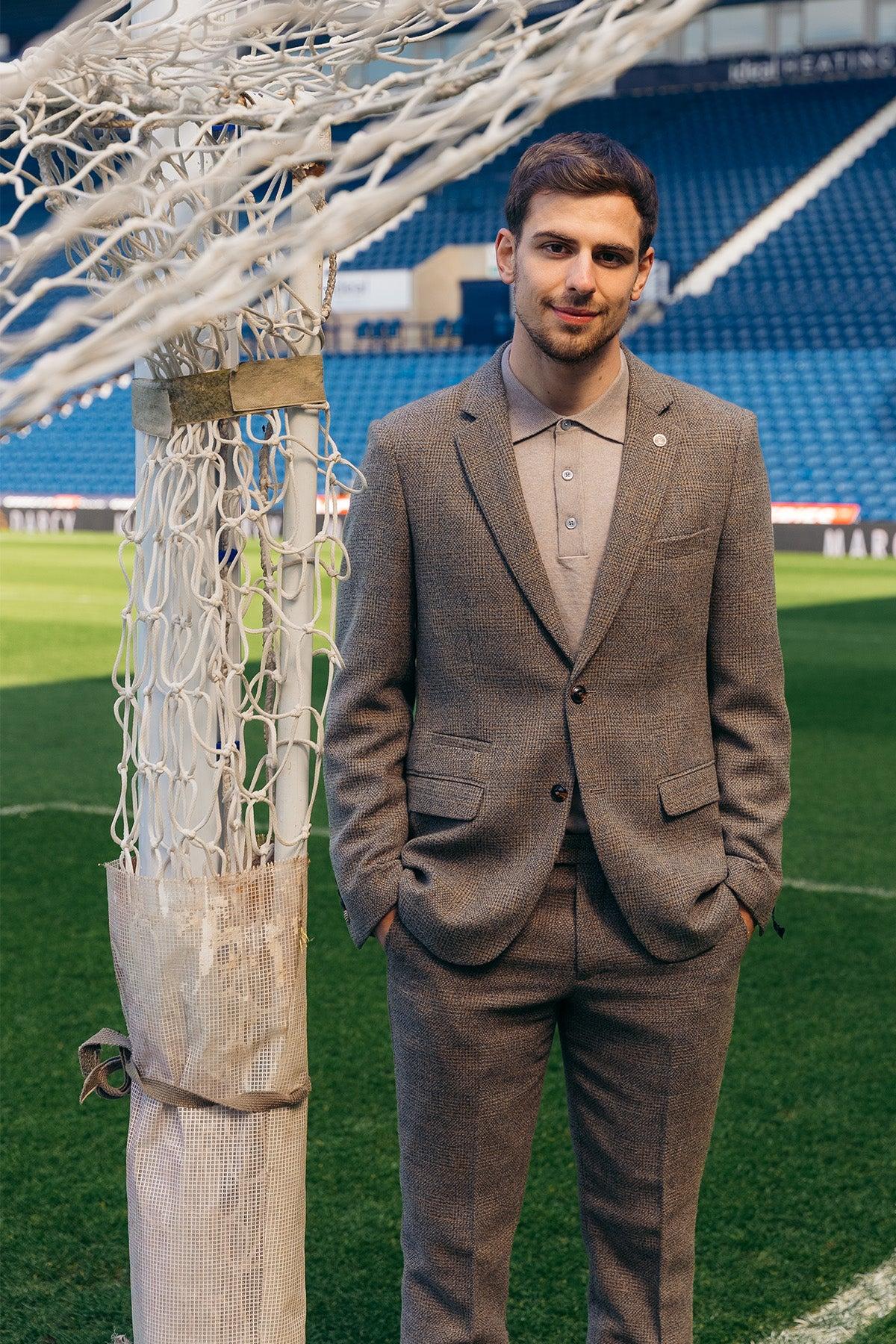 A man in the marcdarcy The West Bromwich Albion F.C. Collection KRIS Tan Tweed Suit, as seen on Jayson Molumby, stands with hands in his pockets by a soccer goalpost on a football field with blue stadium seats behind.