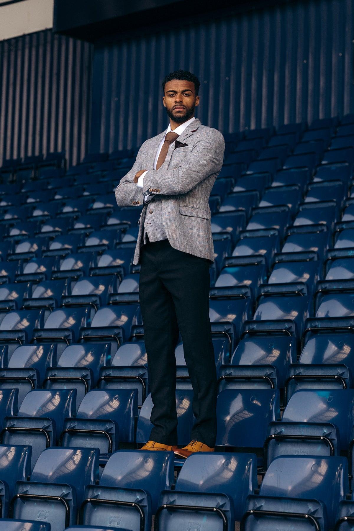 A man wears the marcdarcy West Bromwich Albion F.C. Collection: KRIS Silver Grey Blazer & Max Black Trousers, as seen on Darnell Furlong, standing confidently with arms crossed on empty stadium seats.