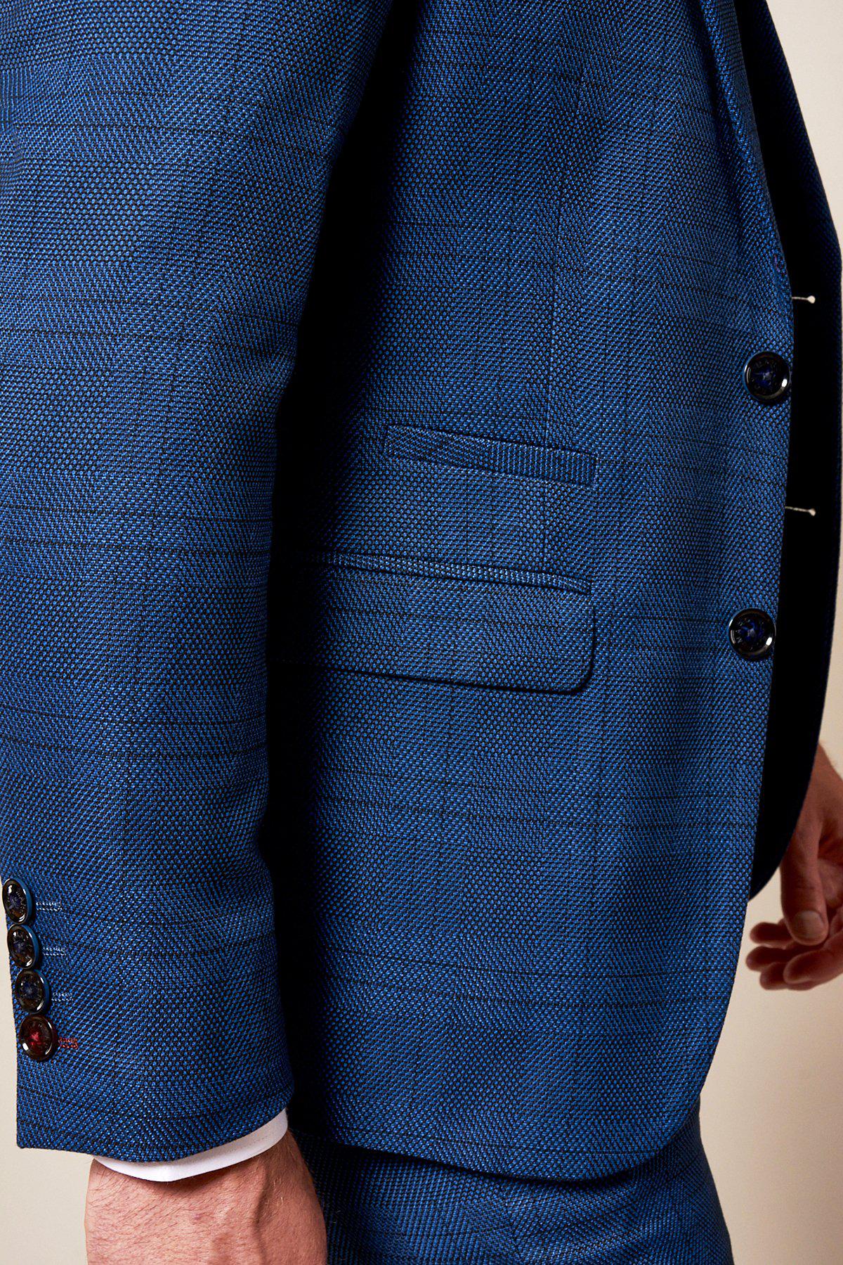 A close-up of a person wearing the marcdarcy WBA Collection JERRY Blue Suit with Jerry Grey Waistcoat, featuring black buttons, a peak lapel, and a front pocket. The hand is partially visible at their side. As worn by Jeremy Sarmiento.