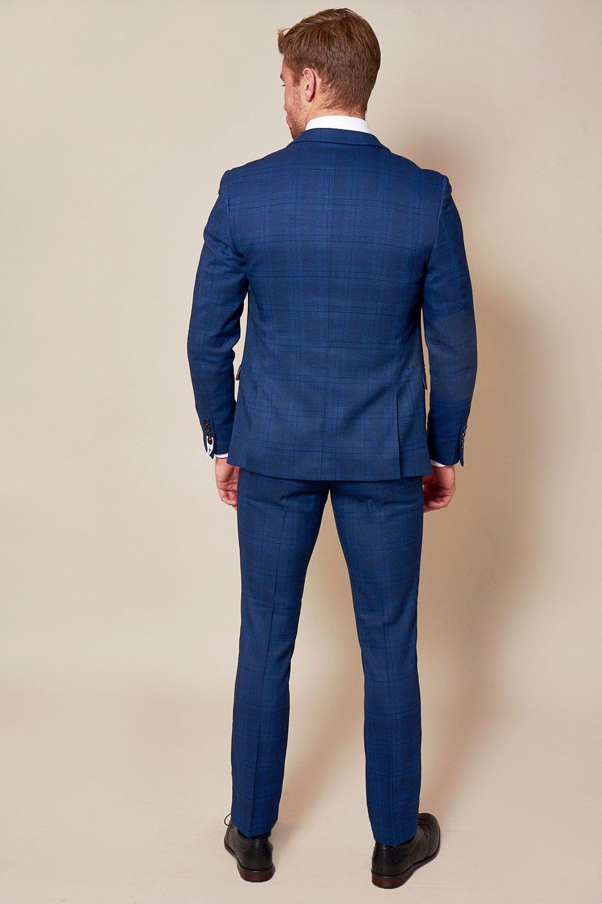 A man faces away from the camera wearing the marcdarcy WBA Collection - JERRY Blue Suit with Jerry Grey Waistcoat as worn by Jeremy Sarmiento, paired with matching trousers, a white shirt, and black dress shoes against a beige background.