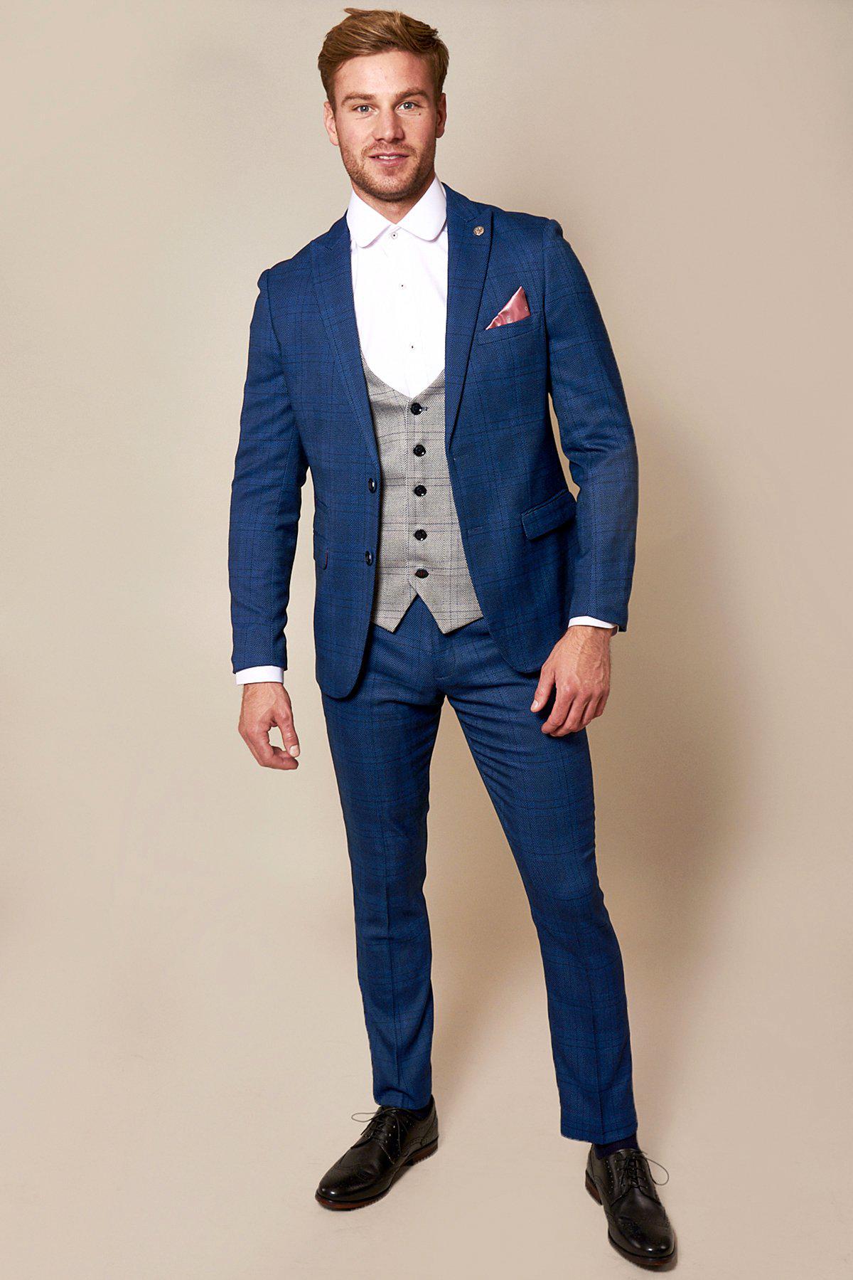 A man models marcdarcy's The WBA Collection - JERRY Blue Suit with Jerry Grey Waistcoat, as worn by Jeremy Sarmiento, paired with a white shirt, pink pocket square, and black shoes.