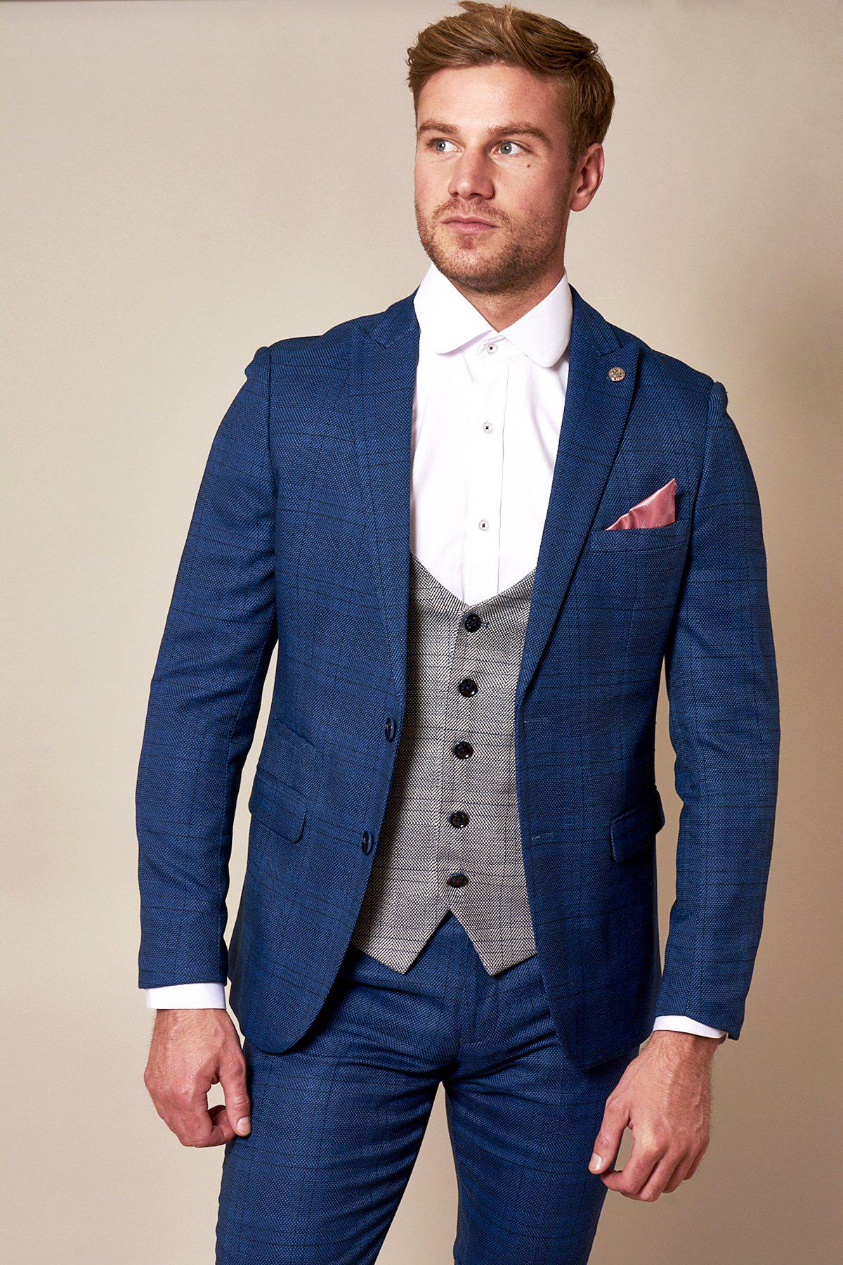A man in the marcdarcy WBA Collection - JERRY Blue Suit with Jerry Grey Waistcoat as worn by Jeremy Sarmiento, paired with a white dress shirt and pink pocket square, stands against a plain light background showcasing sharp, modern style.