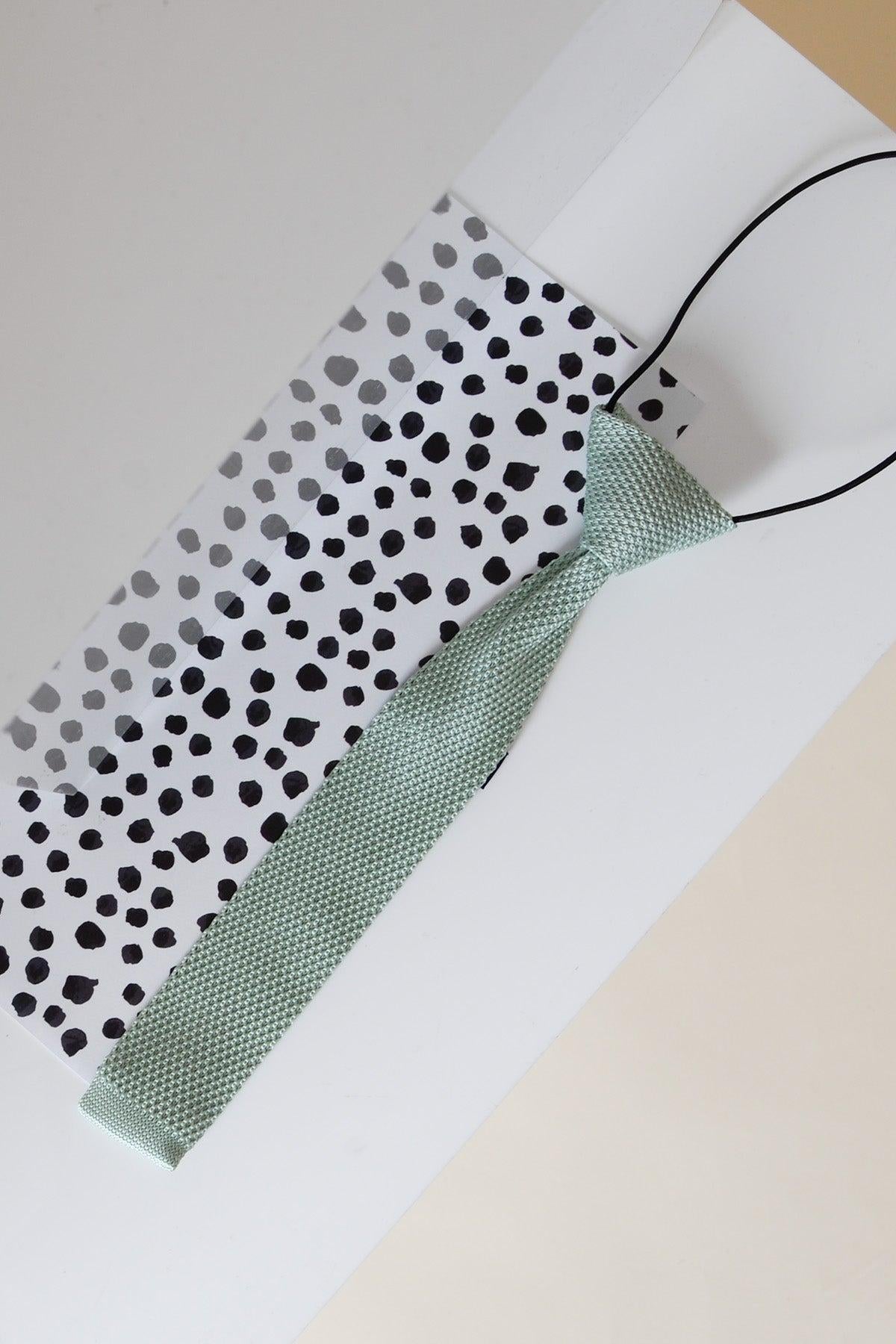 The Marc Darcy Children's Knitted Tie In Green rests on a white and black polka dot gift bag, both displayed on a white surface.