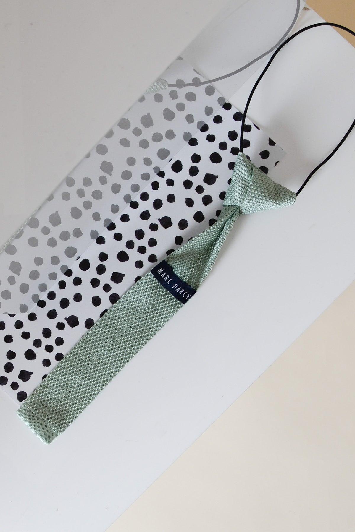The Marc Darcy Children's Knitted Tie in Green is displayed flat on a white surface beside a black and white polka dot accessory, making it the perfect addition to a children's three piece suit.
