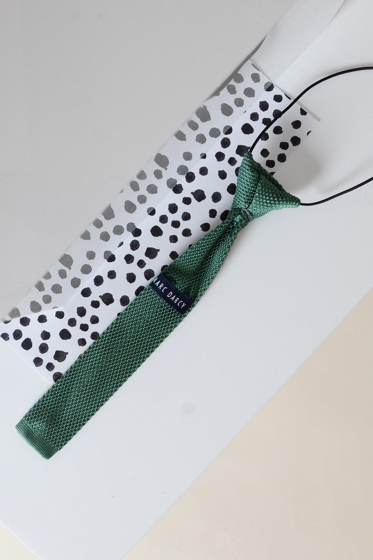 A Marc Darcy Children's Knitted Tie in olive rests on a white surface next to a white gift bag with black polka dots and a black string handle.