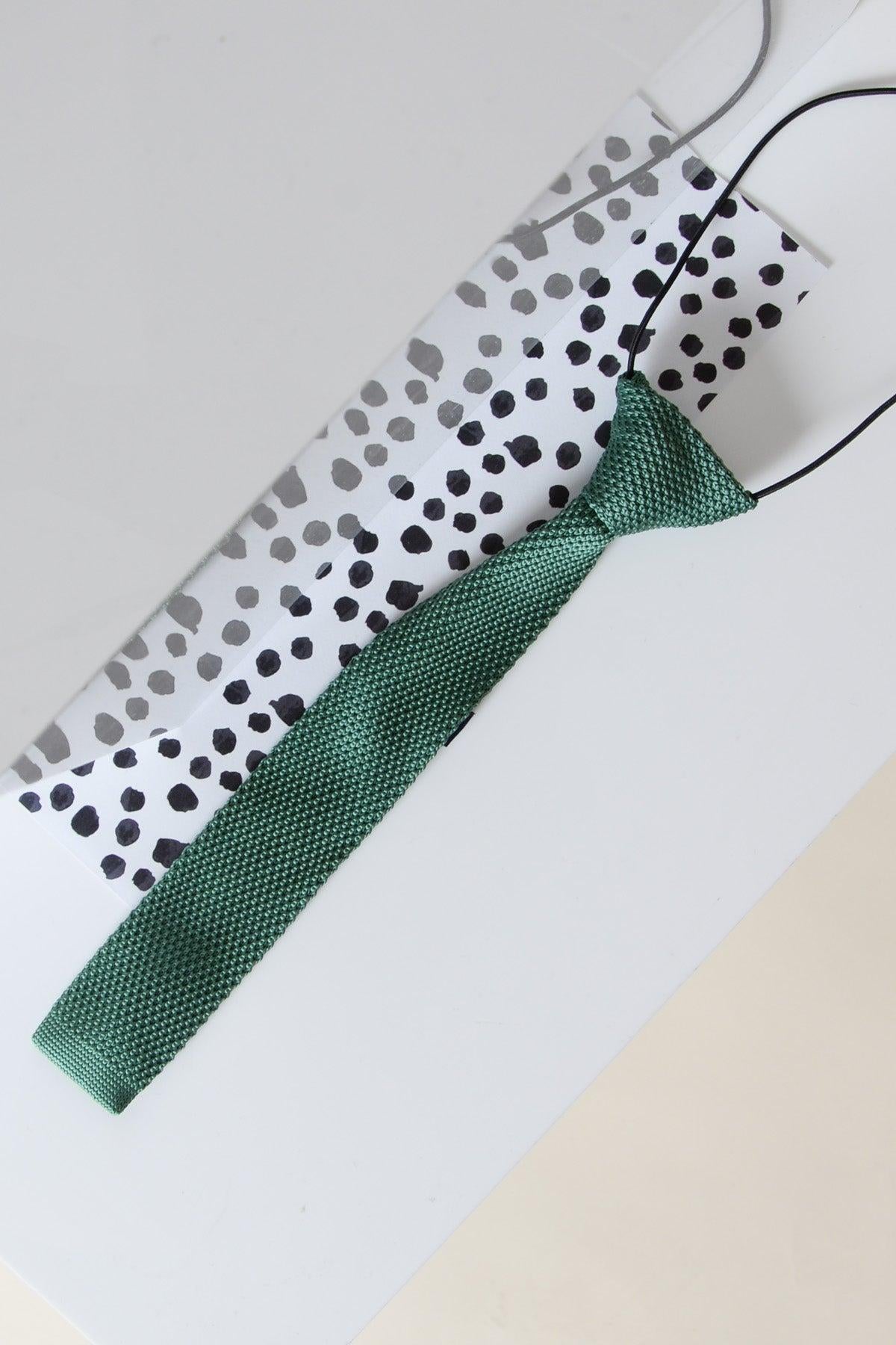 The Marc Darcy Children's Knitted Tie In Olive, featuring a textured knit and elastic band, rests on a white surface beside a dotted white envelope.