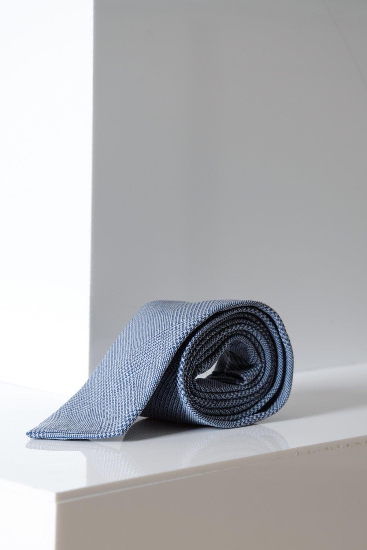 The BROMLEY - Sky Check Tie by Accessory, featuring a light blue Prince of Wales check design, is rolled up and displayed on a glossy white surface with a minimalist, light background.