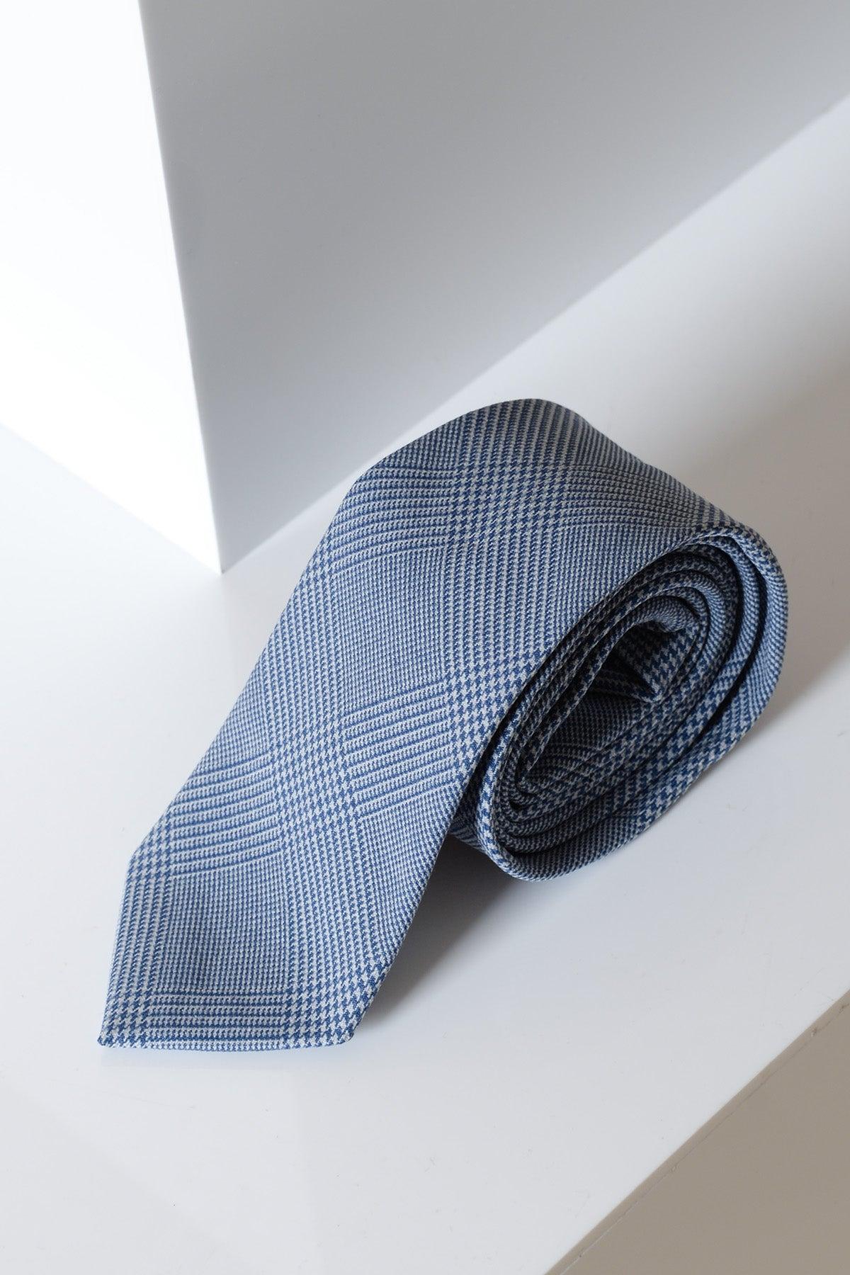 The BROMLEY - Sky Check Tie by Accessory features a blue check pattern, neatly rolled on a white geometric surface with soft lighting—ideal for elevating your formalwear look.