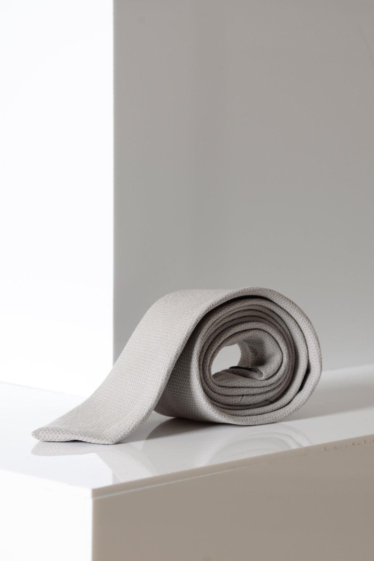 The HM5 - Stone Tie by Accessory, neatly rolled in light gray, rests on a glossy white surface against a minimalist white background.