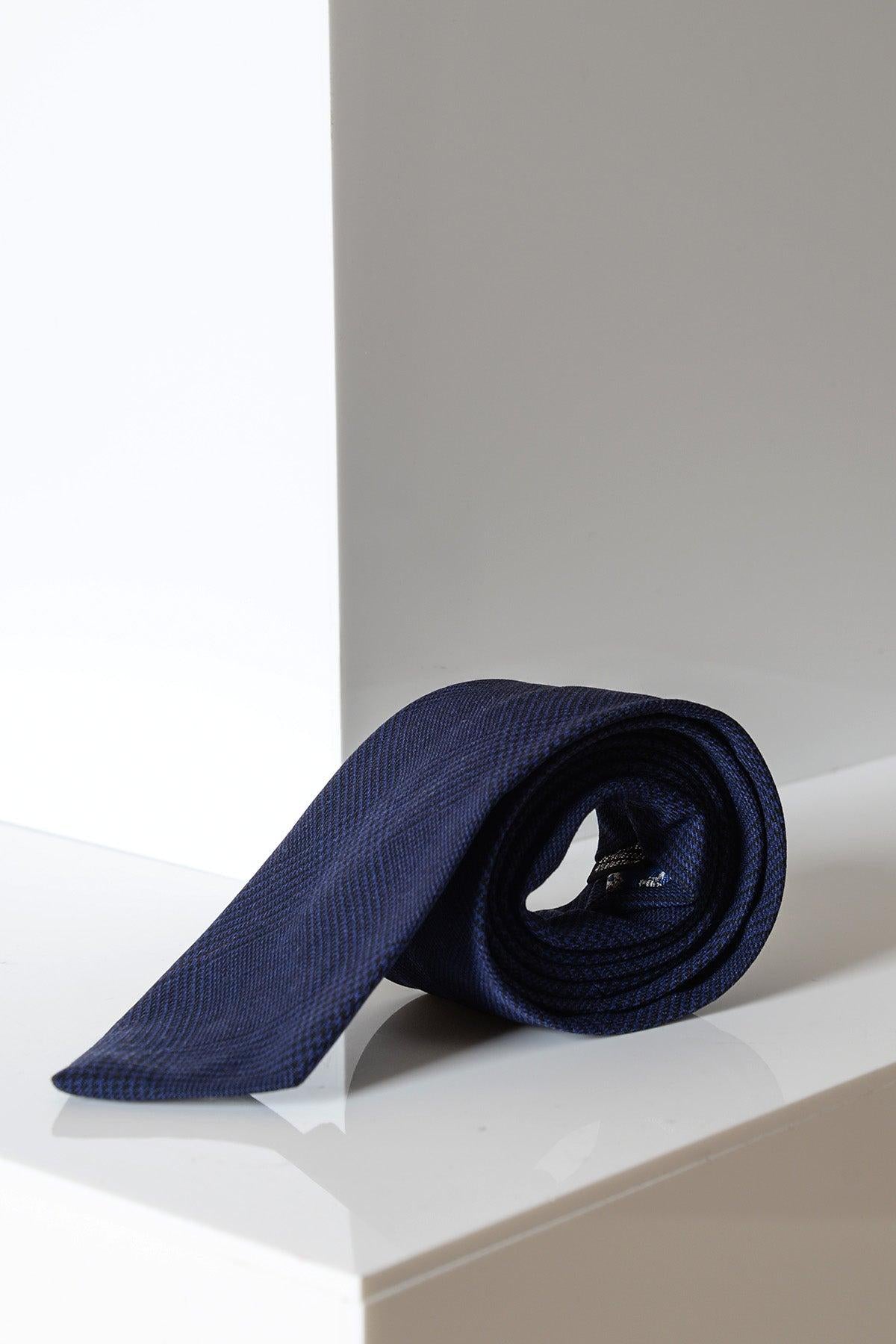 The BROMLEY - Navy Check Tie by Accessory, featuring a subtle diamond pattern, is handmade and displayed rolled on a white surface.