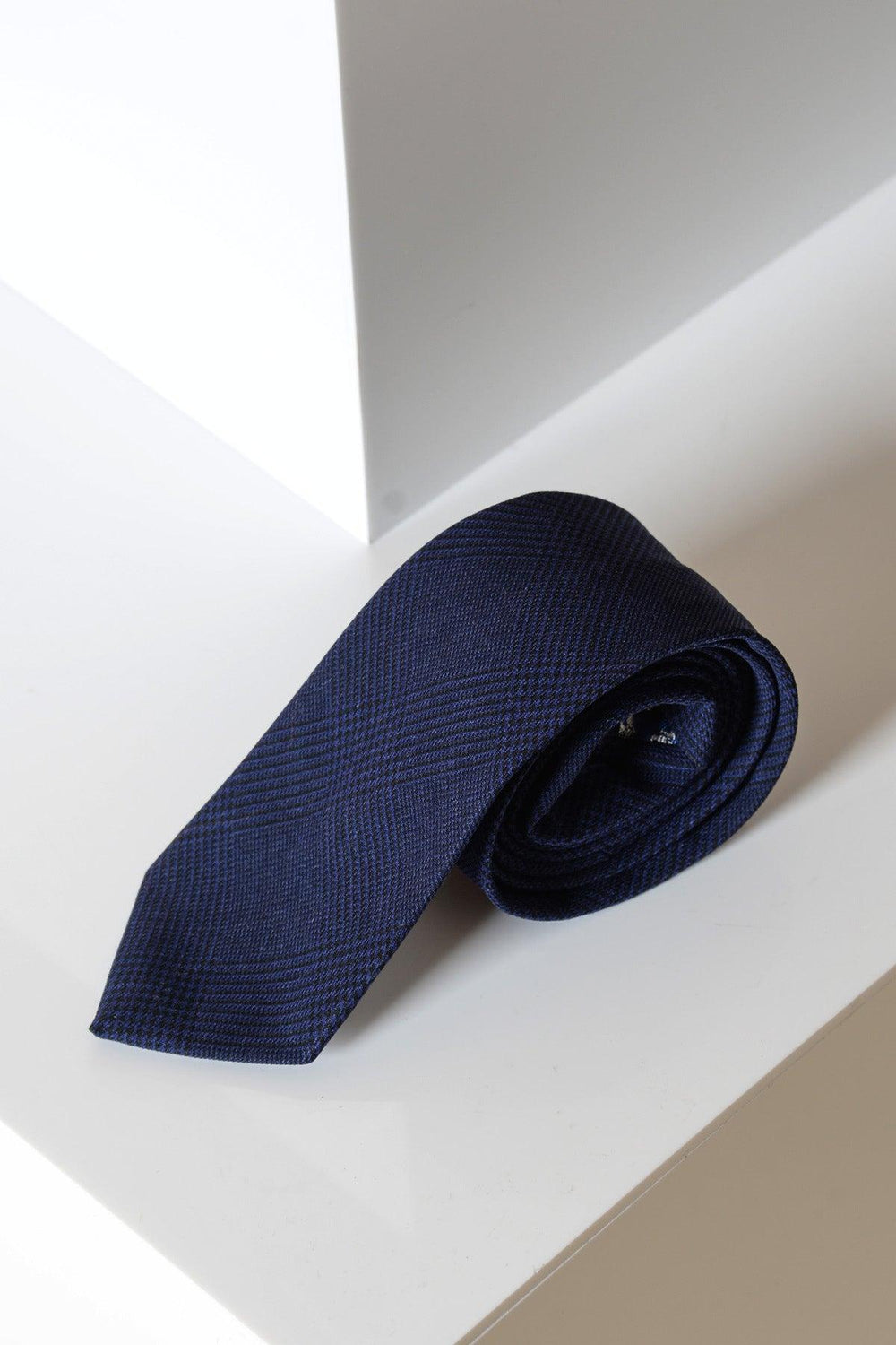 The BROMLEY - Navy Check Tie by Accessory, featuring a handmade check pattern, is rolled up on a glossy white surface near the corner of a white geometric structure.