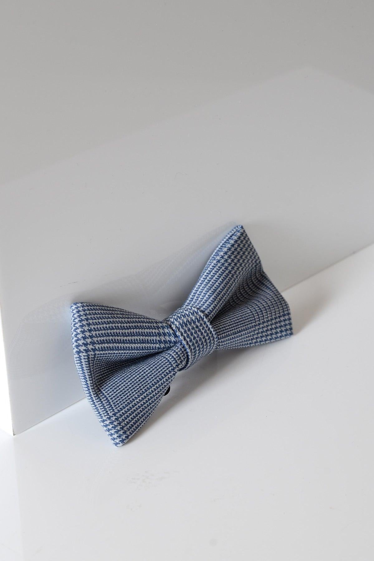 The BROMLEY - Sky Check Bow Tie by Accessory features a light blue and white checkered pattern, elegantly displayed against a white reflective surface for a minimal, refined formalwear look.
