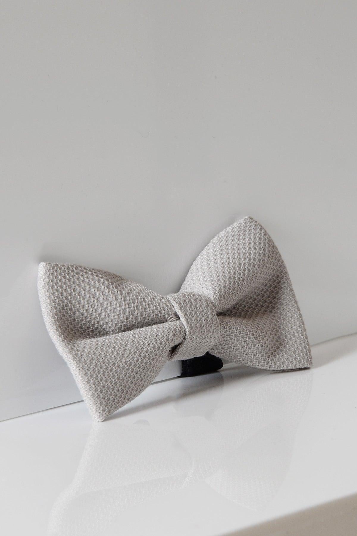 The HM5 - Stone Bow Tie by Accessory features a textured light gray design, displayed on a white reflective surface. This refined piece is an ideal wedding accessory and the perfect addition to any formalwear look.