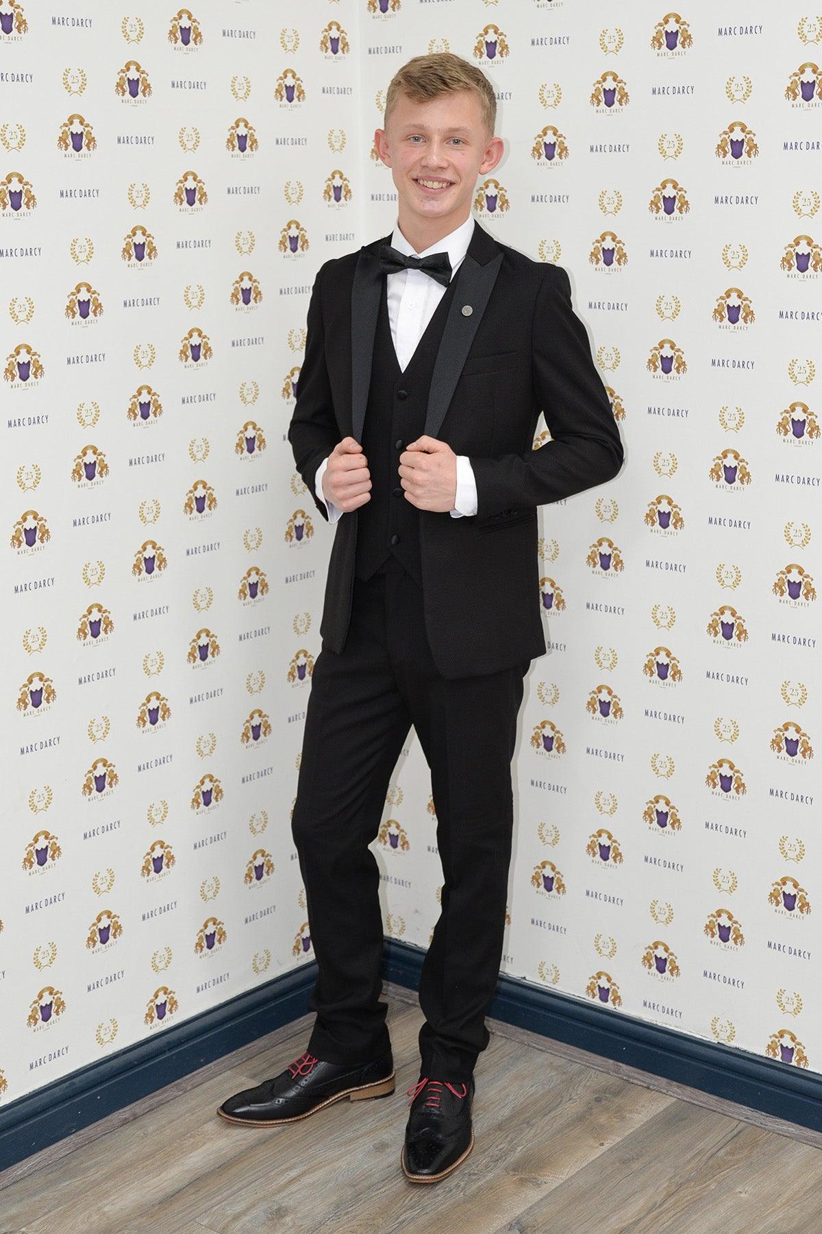 Coronation Street's Dylan Wilson (Liam McCheyne) wears the Dalton Black Tuxedo Suit by marcdarcy, standing before a wall with the marcdarcy logo, ready for any black-tie event.
