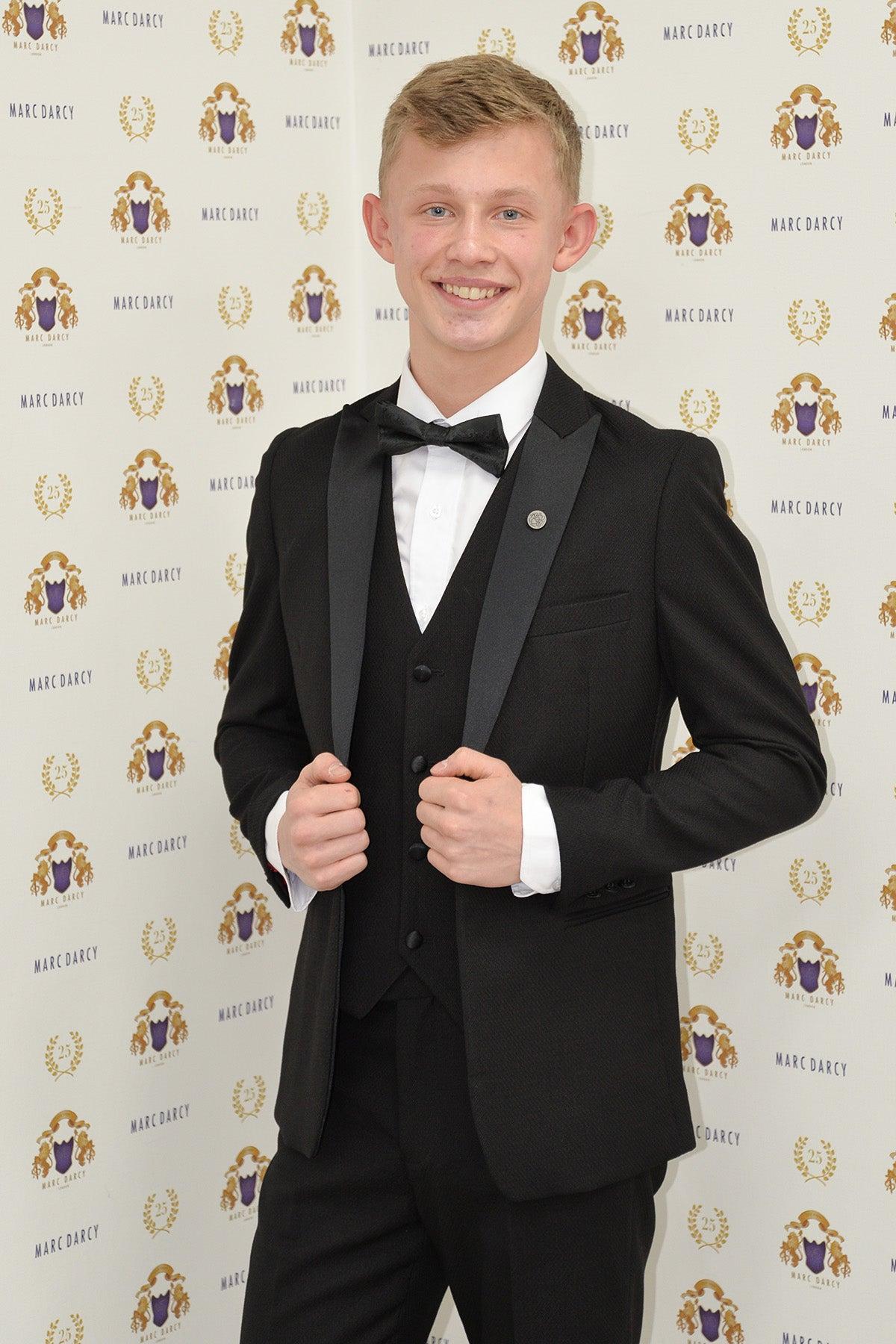 Coronation Street’s Dylan Wilson (Liam McCheyne) wears the marcdarcy Dalton Black Tuxedo Suit with satin peak lapel, smiling and holding his jacket in front of a Marc Darcy logo backdrop.