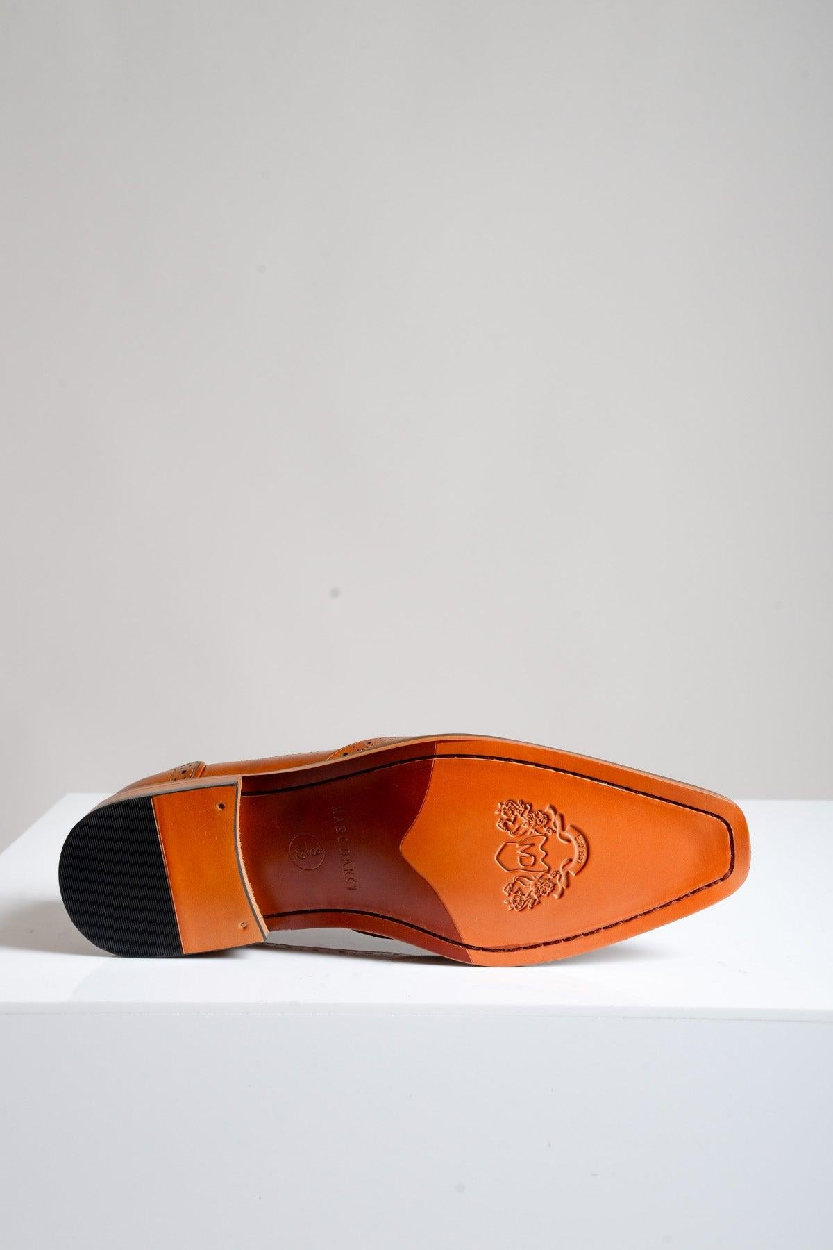 The Marc Darcy LARKIN - Tan Leather Brogue Shoe is displayed upside down, showcasing its smooth tan sole with hand-stitched edging and an embossed lion head. The shoe features a black heel and is set against a plain light background.