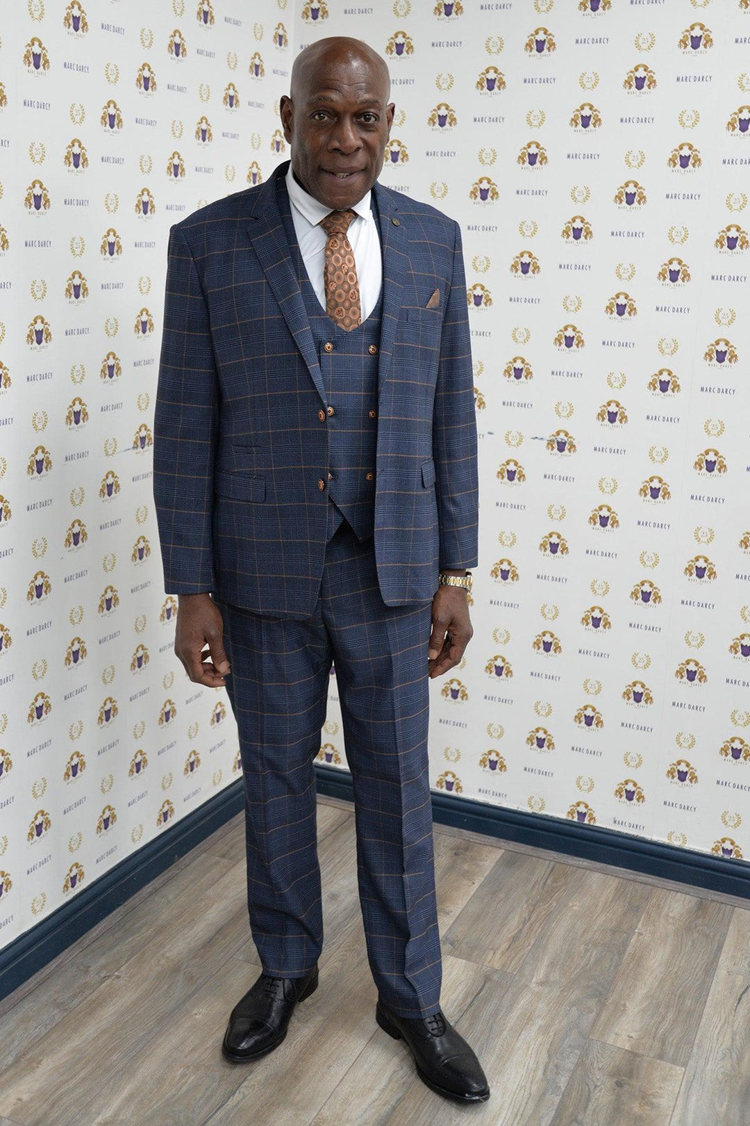 Boxing legend Frank Bruno wears the marcdarcy Jenson Marine Check Suit, paired with a white shirt and patterned tie, standing on a wood floor before a white wall with multicolored logos, smiling with his hands by his sides.