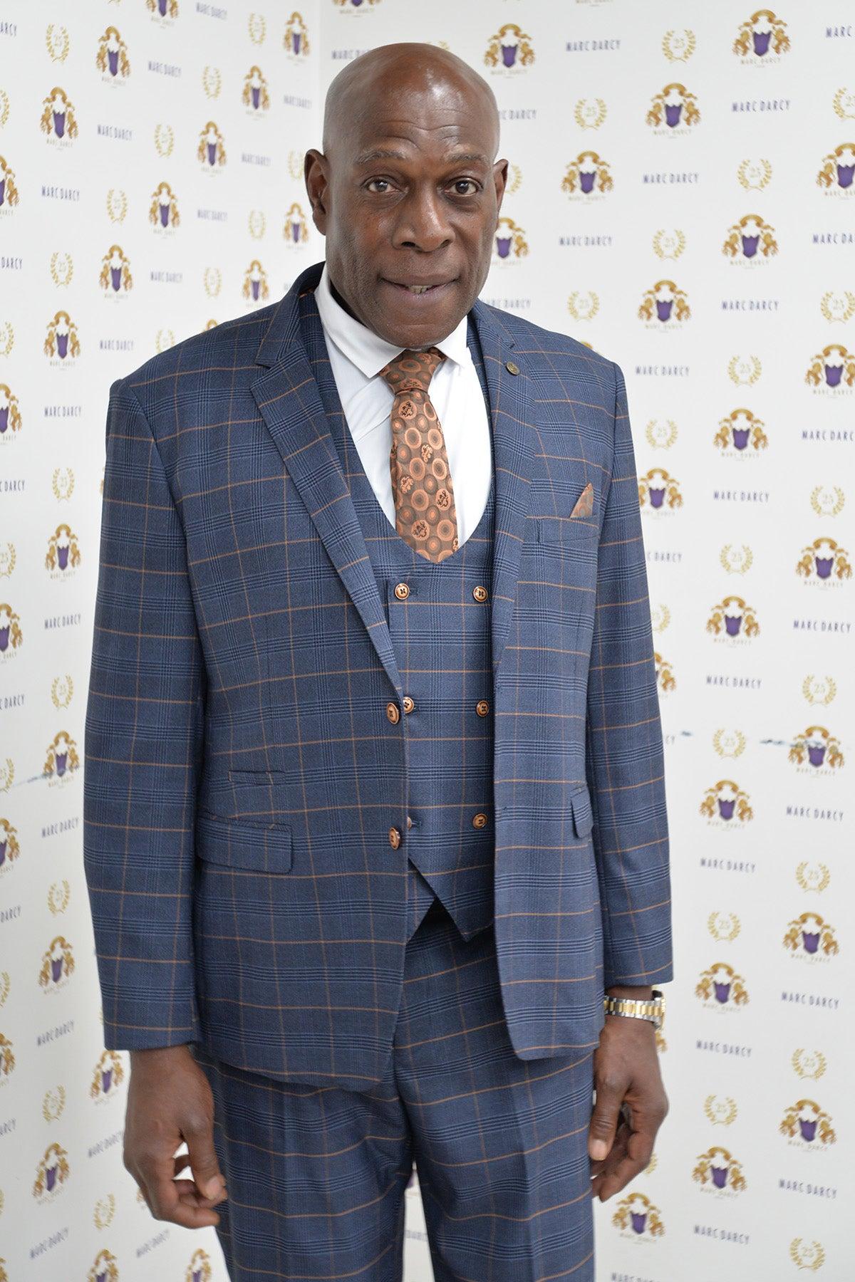 Boxing legend Frank Bruno wears a marcdarcy Jenson Marine Check suit, featuring a double-breasted waistcoat and patterned tie, as he stands before a white backdrop with gold and blue logos.