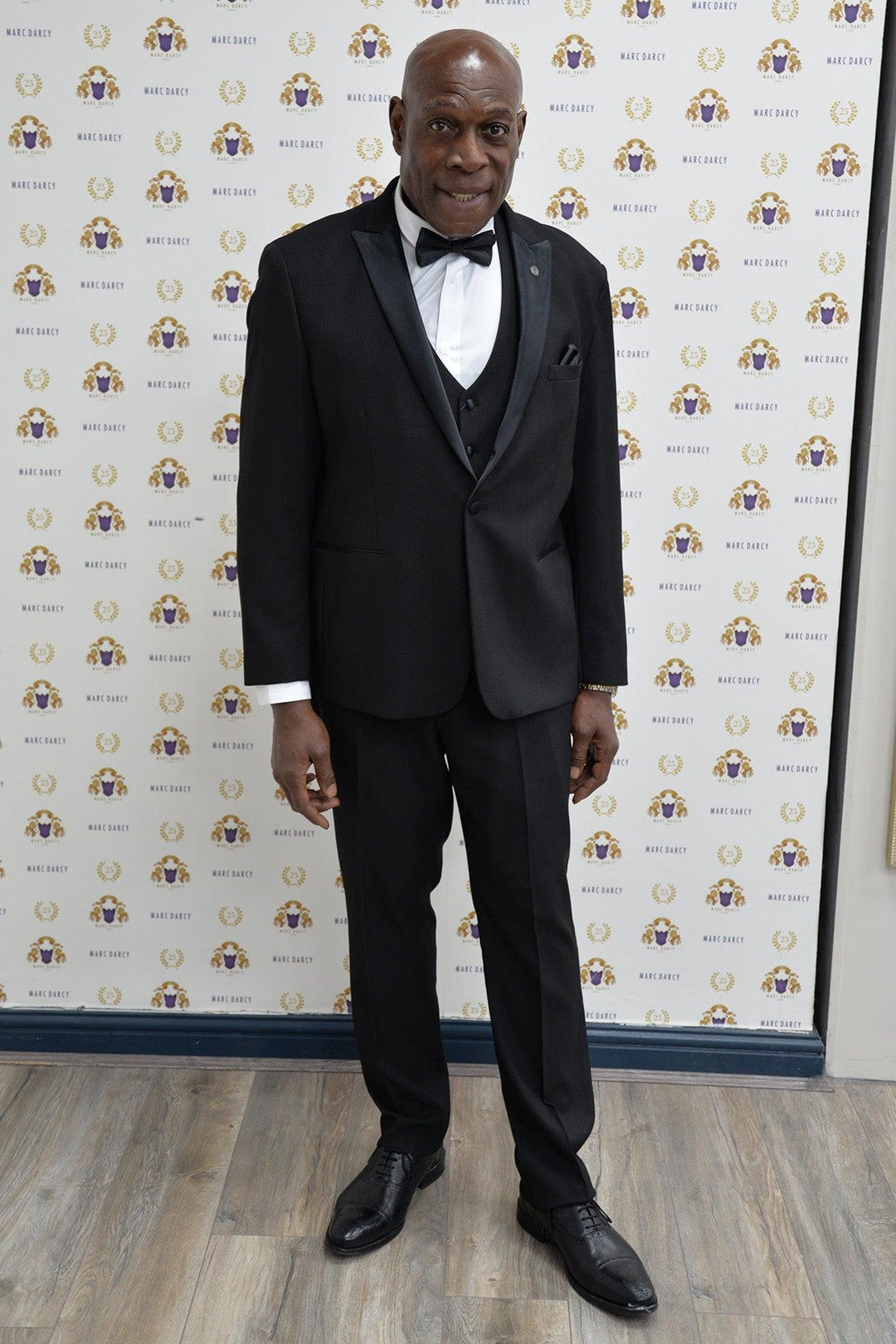 Boxing legend Frank Bruno wears the marcdarcy DALTON Black Tux Lapel Suit, standing on a wooden floor before a white backdrop with gold and purple logos, smiling confidently at the camera.