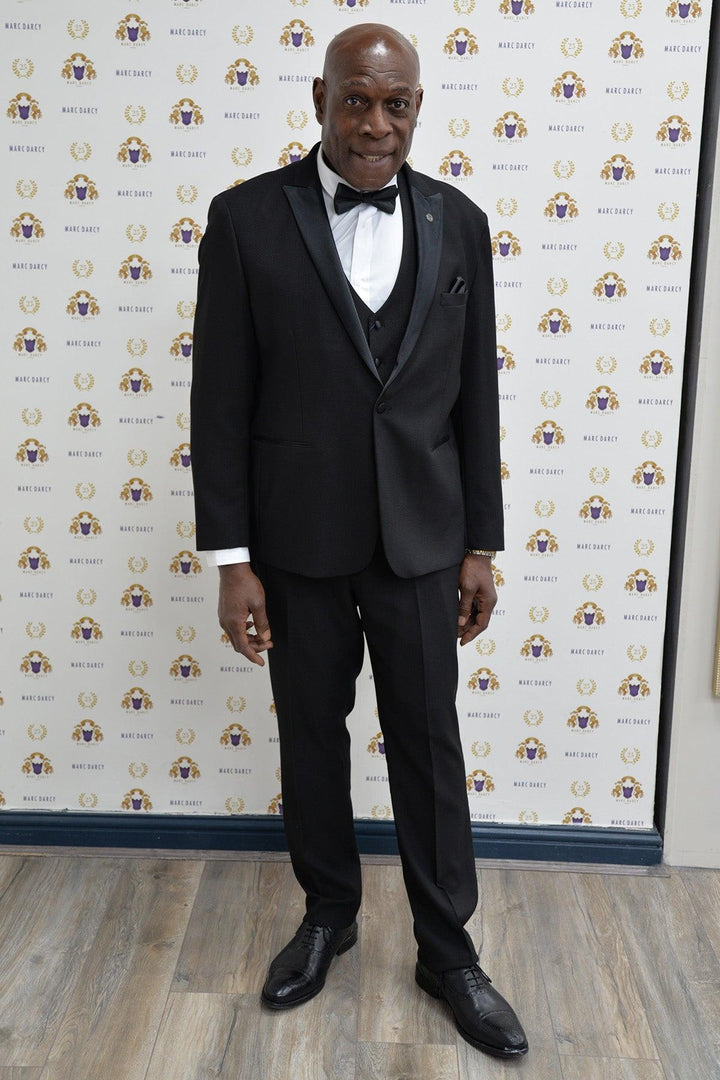 Boxing legend Frank Bruno wears the marcdarcy DALTON Black Tux Lapel Suit, standing on a wooden floor before a white backdrop with gold and purple logos, smiling confidently at the camera.